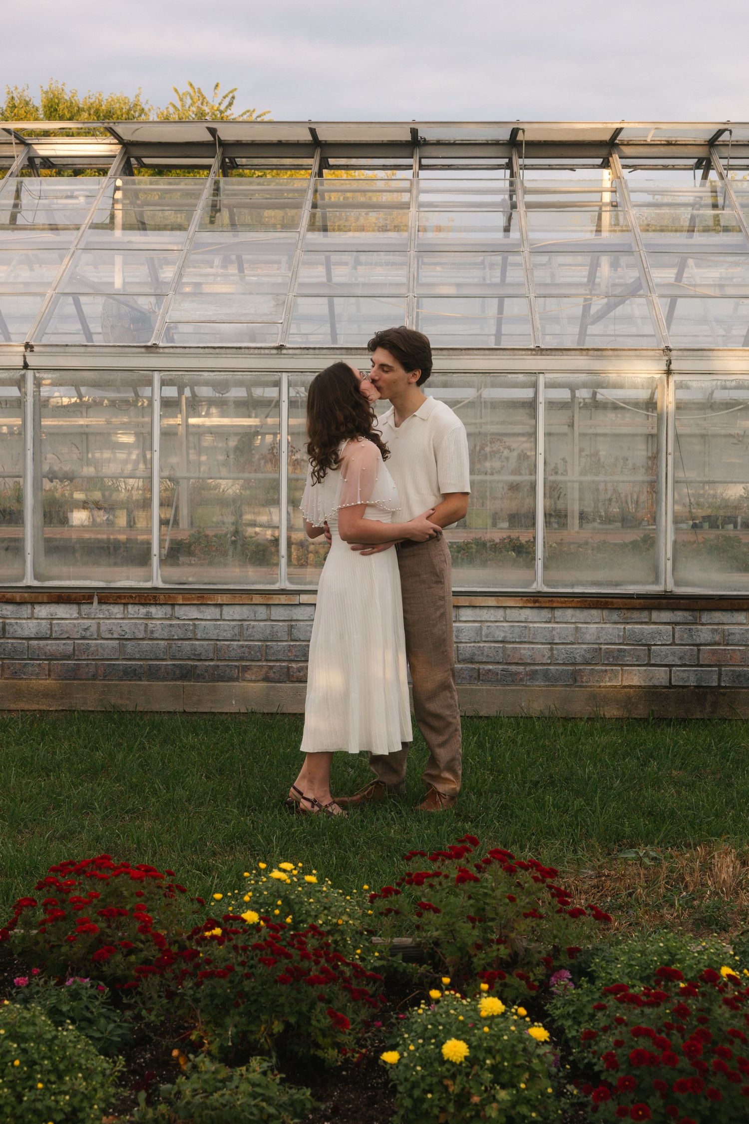 Couple embracing and kissing in front of a glass greenhouse surrounded by colorful red and yellow flowers.