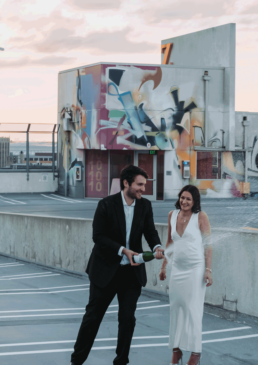 Couple celebrating on rooftop with champagne, wearing wedding attire, colorful graffiti mural backdrop.