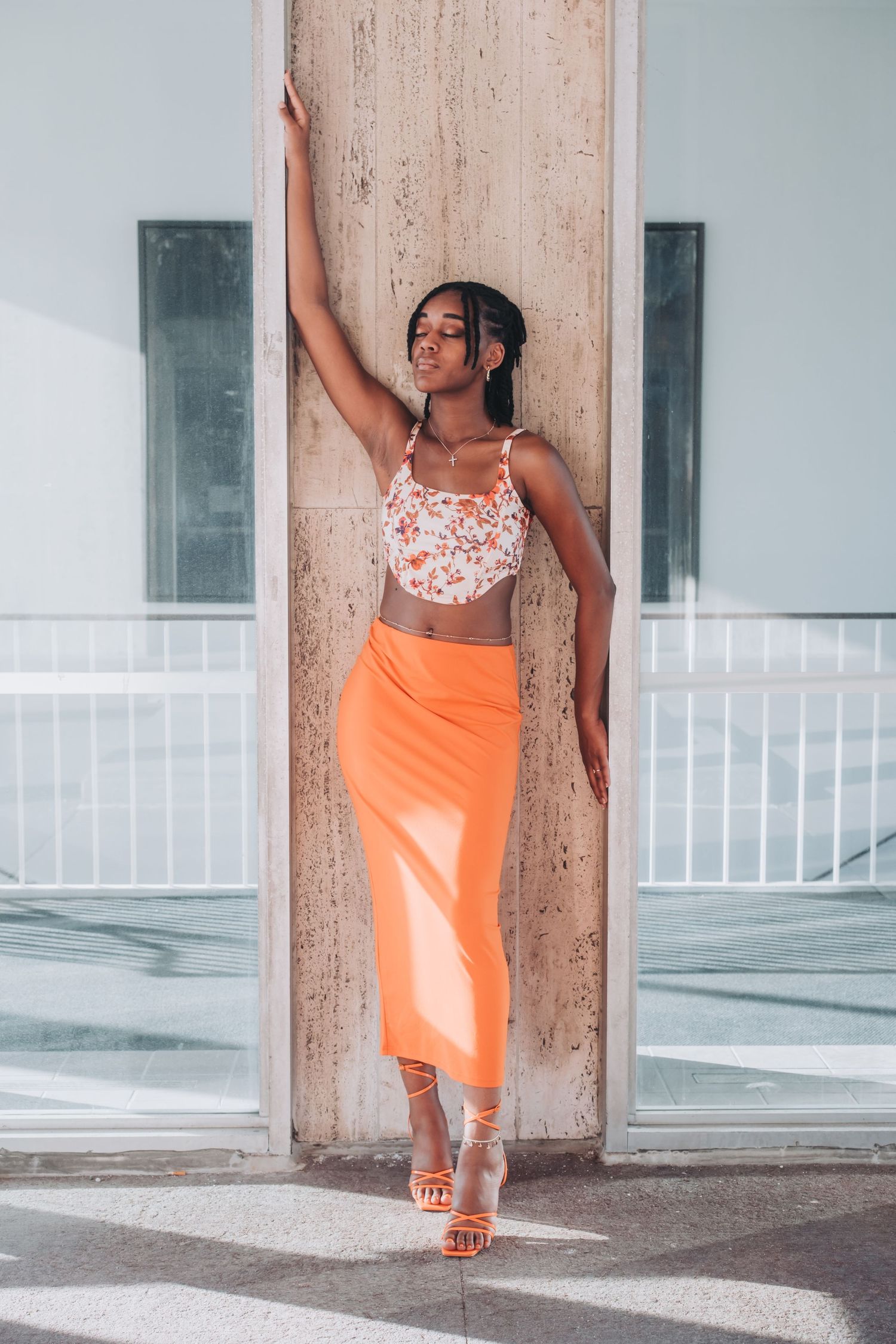 Young woman in floral crop top and orange midi skirt poses against a concrete pillar in heeled sandals.