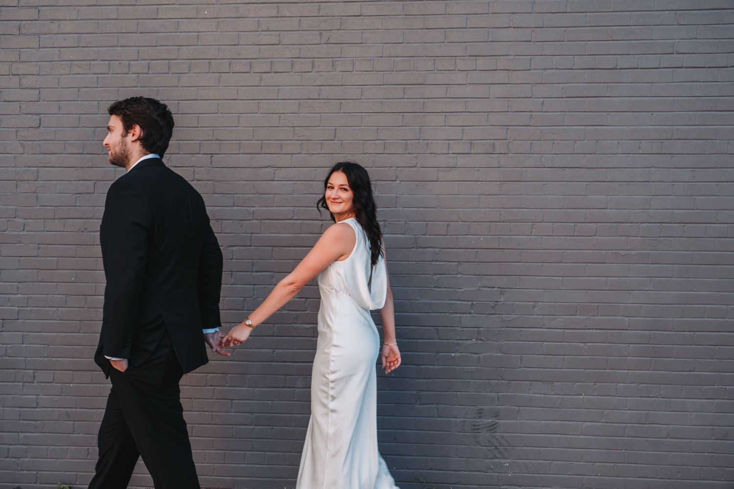 Bride in white gown and groom in black tuxedo walk hand-in-hand past a gray brick wall.