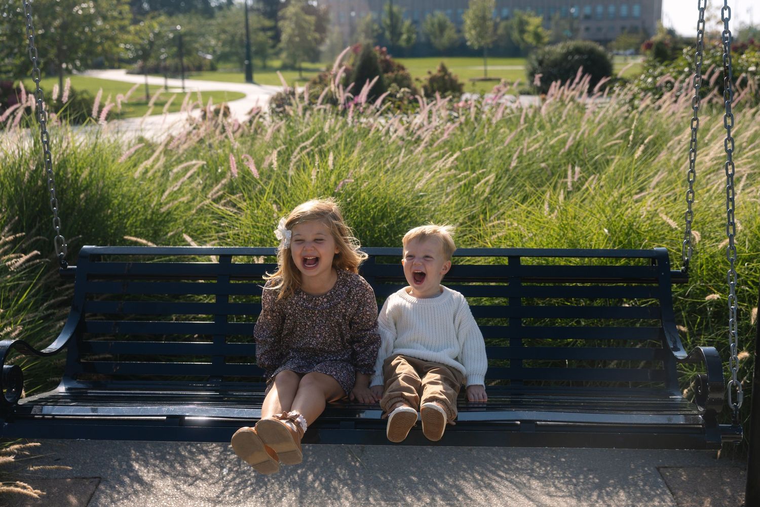 Two children sit together on a green park bench on a sunny day with tall grasses swaying in the background.