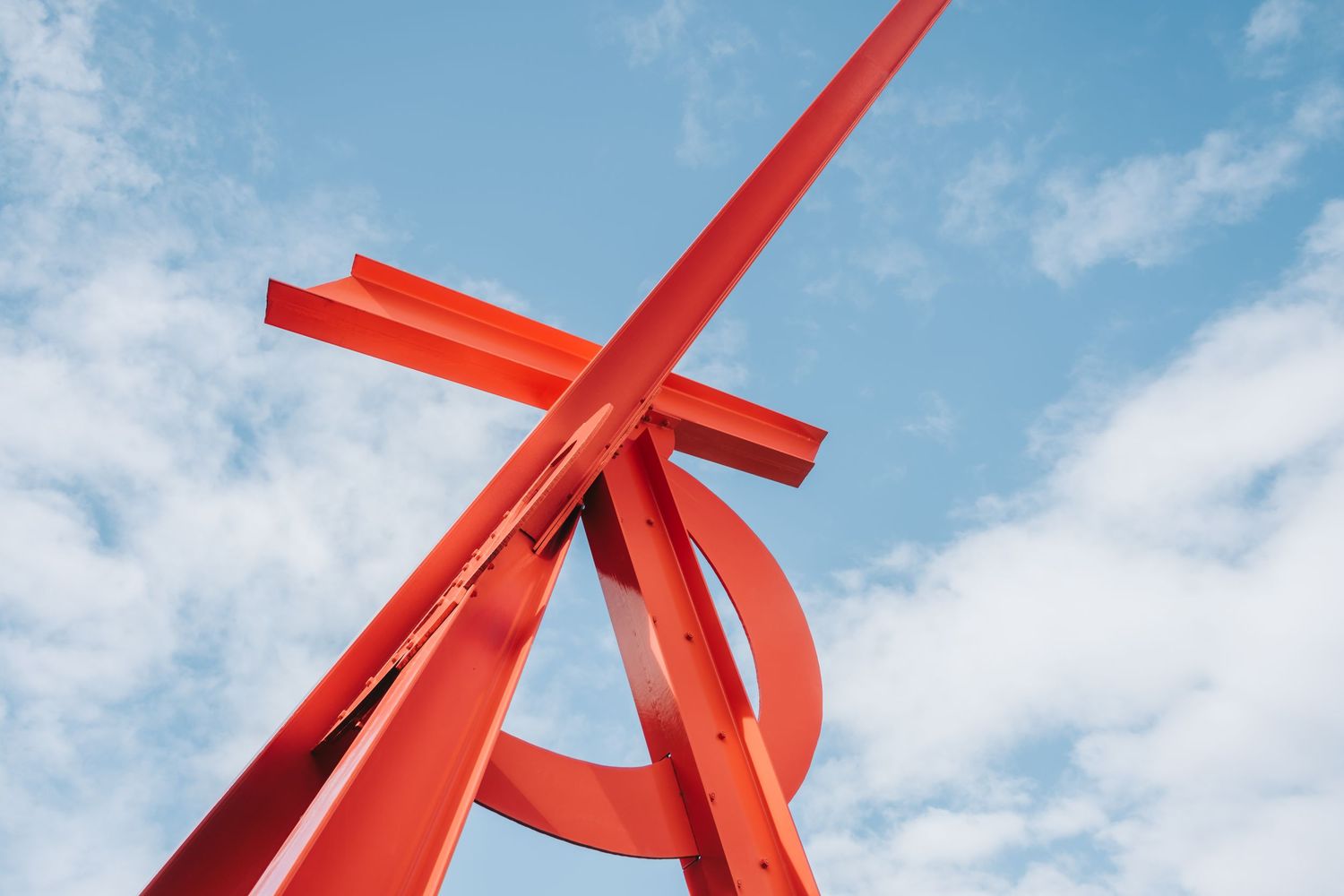 A bold red abstract metal sculpture against a blue sky with white clouds, featuring intersecting geometric steel beams.