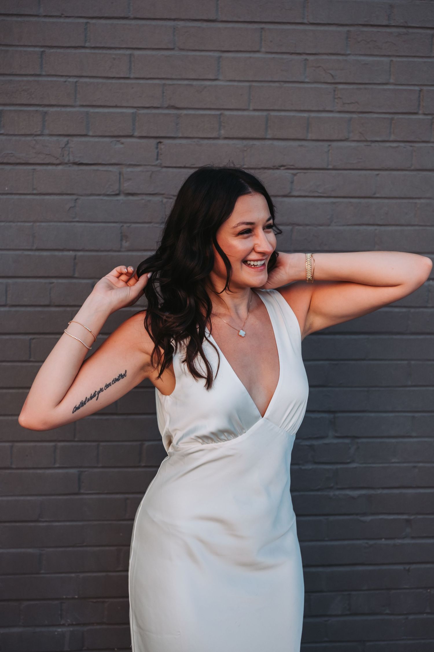 Smiling brunette woman in a white deep V-neck dress with arm tattoo posing against a gray brick wall.