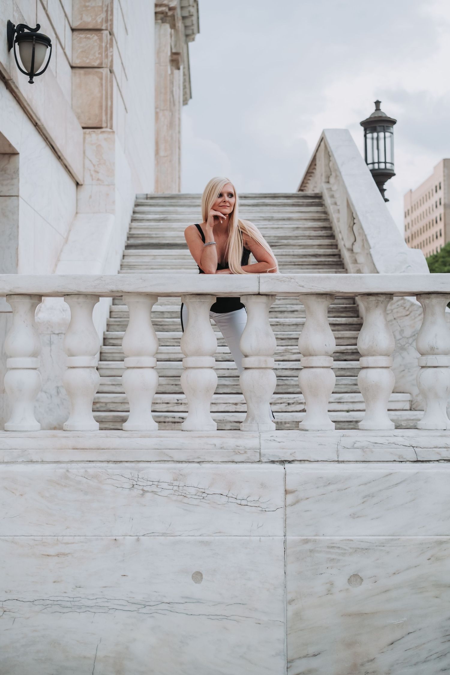 Blonde woman leaning on white marble balustrade staircase of elegant building, smiling outdoors.