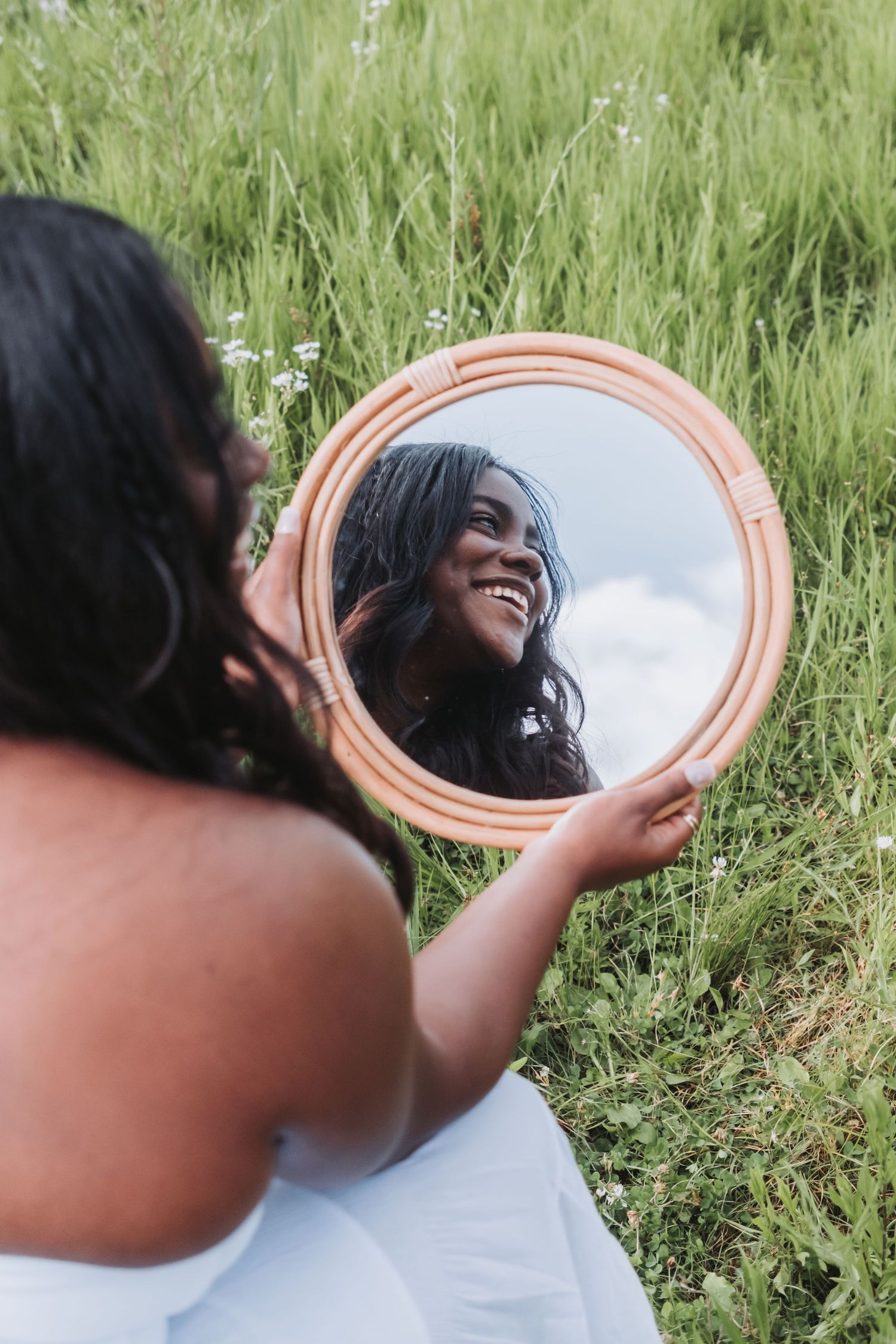 A smiling Black woman in a white dress holds a round mirror reflecting her joyful face while sitting in a lush green field.