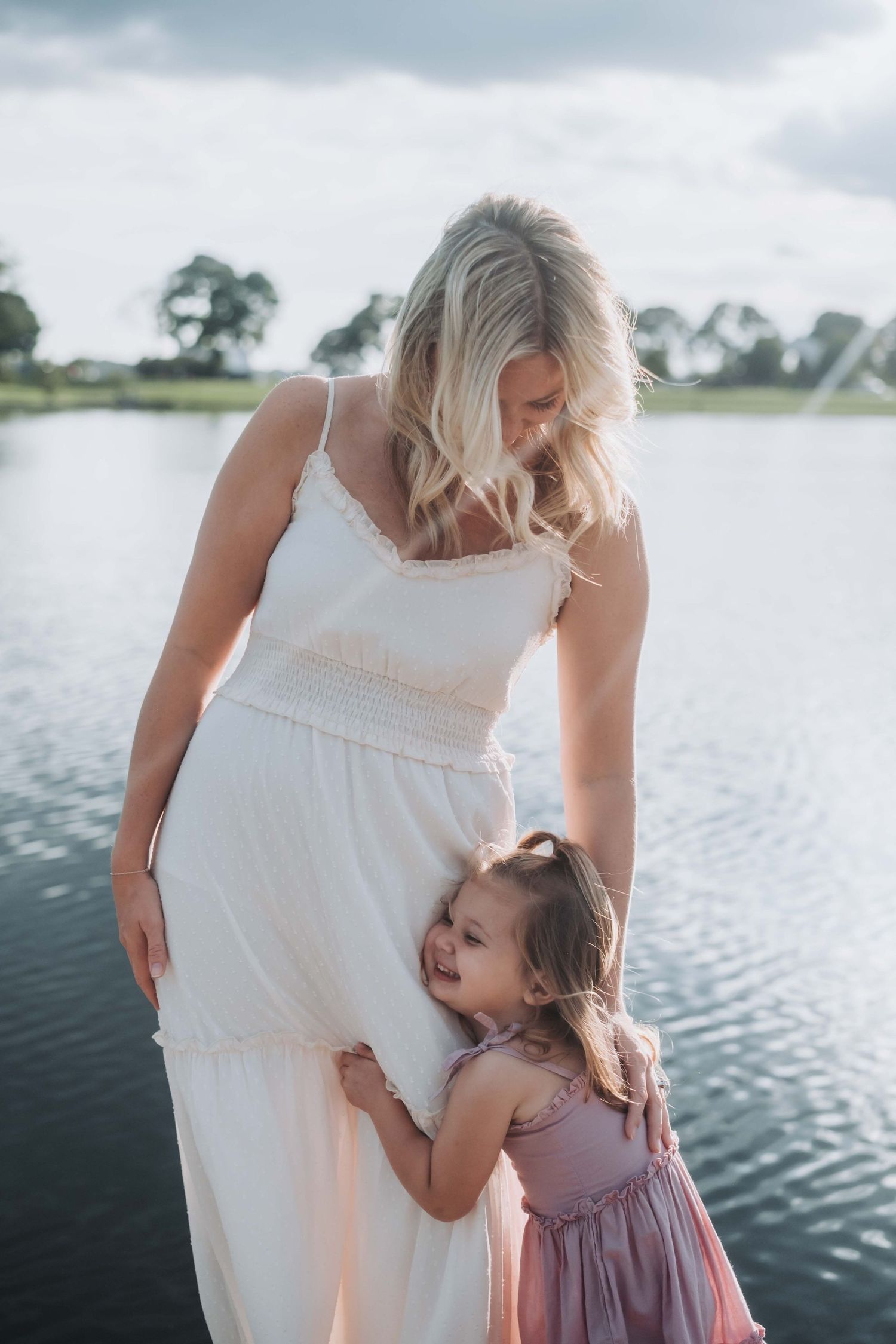 A blonde woman in a white dress hugs her young daughter in a pink dress near a serene lake outdoors.