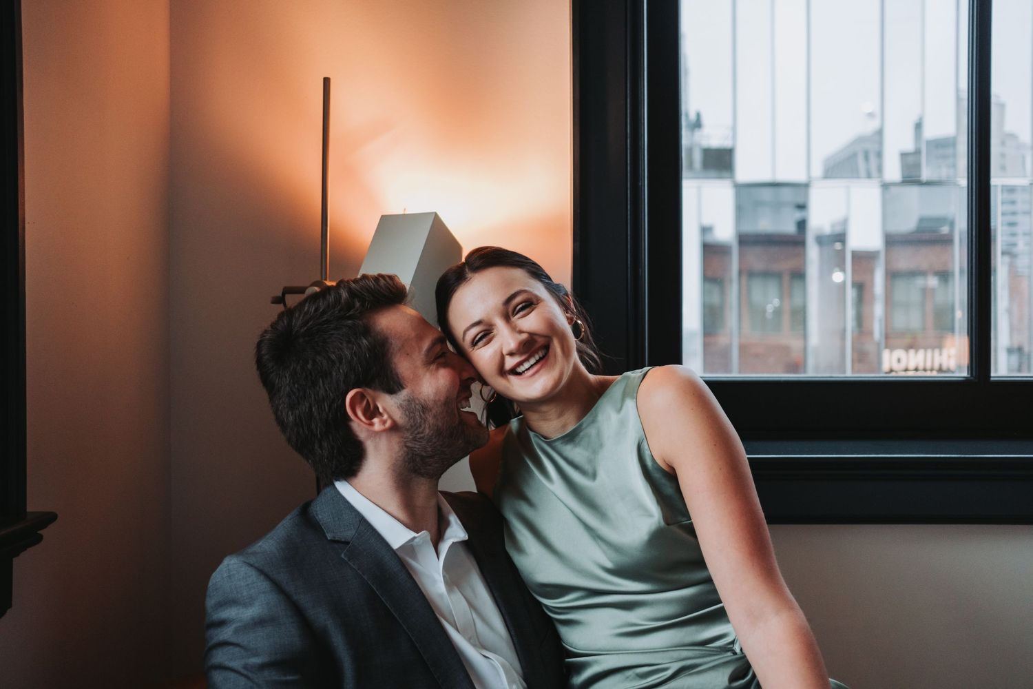 A smiling couple shares a romantic moment, man in gray suit whispering to laughing woman in green dress by a window.
