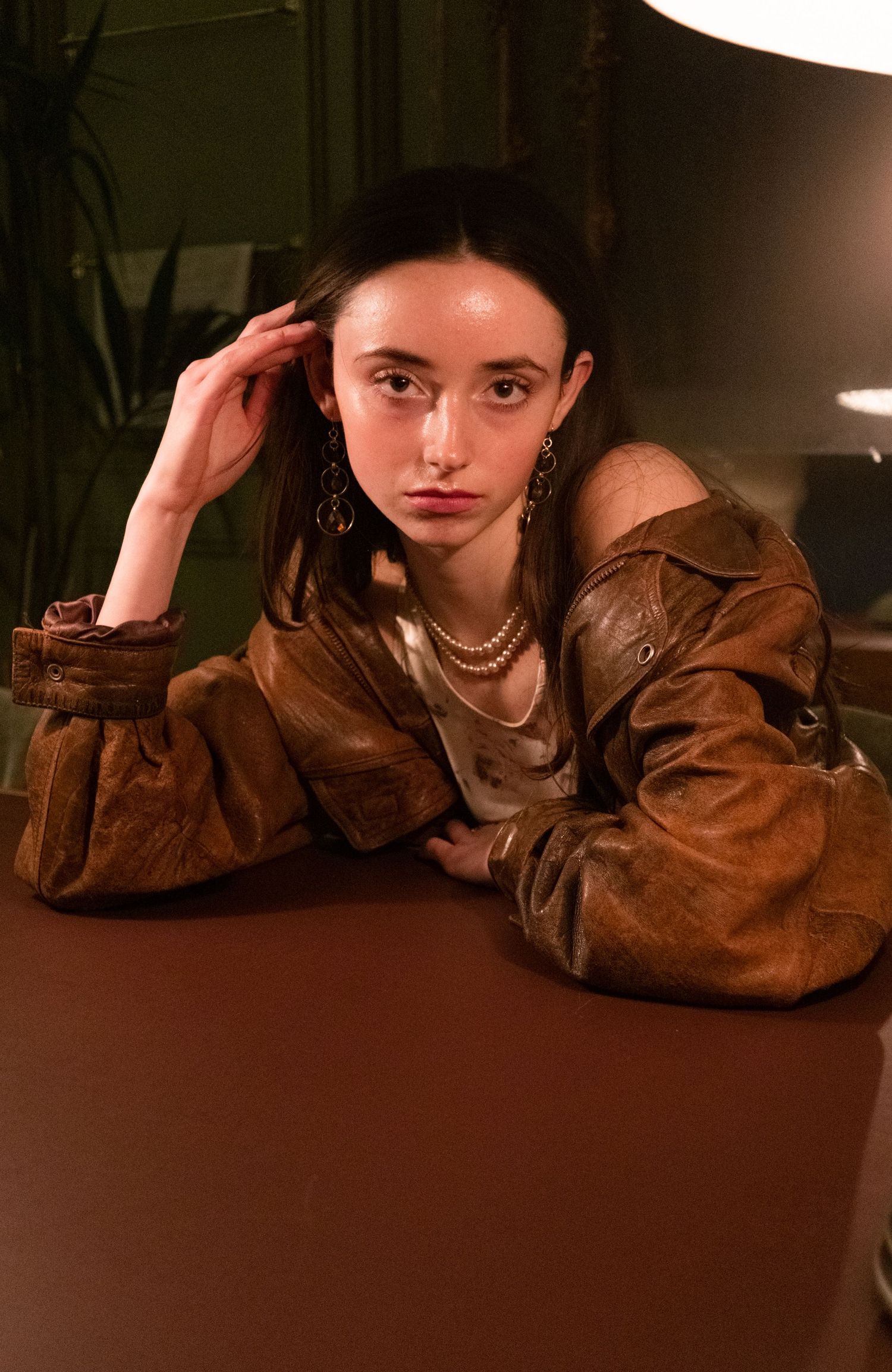 Woman in vintage brown leather jacket with gold jewelry poses at table.