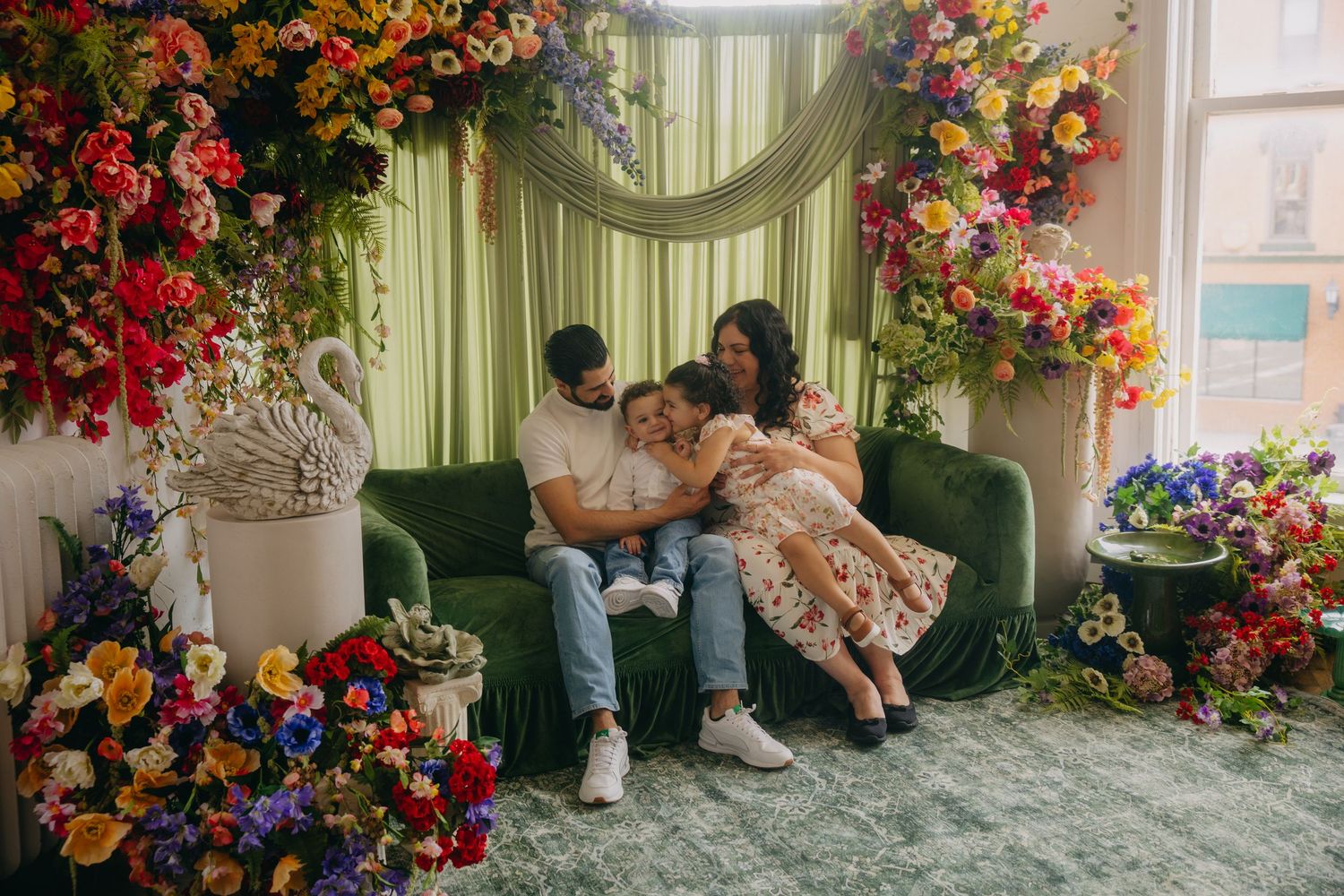 Family of four sitting on green velvet sofa surrounded by colorful floral arrangements in elegant studio setting.