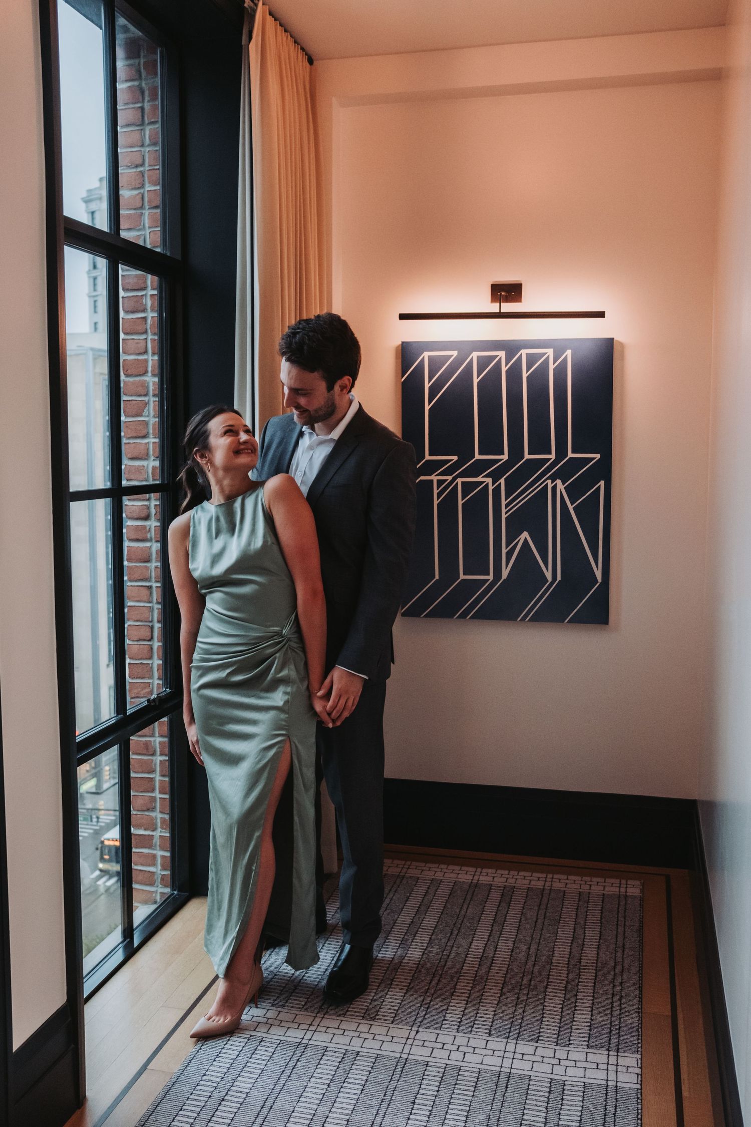 Couple in formal attire embracing by a window near a Pool Town art print in an elegant hotel corridor.