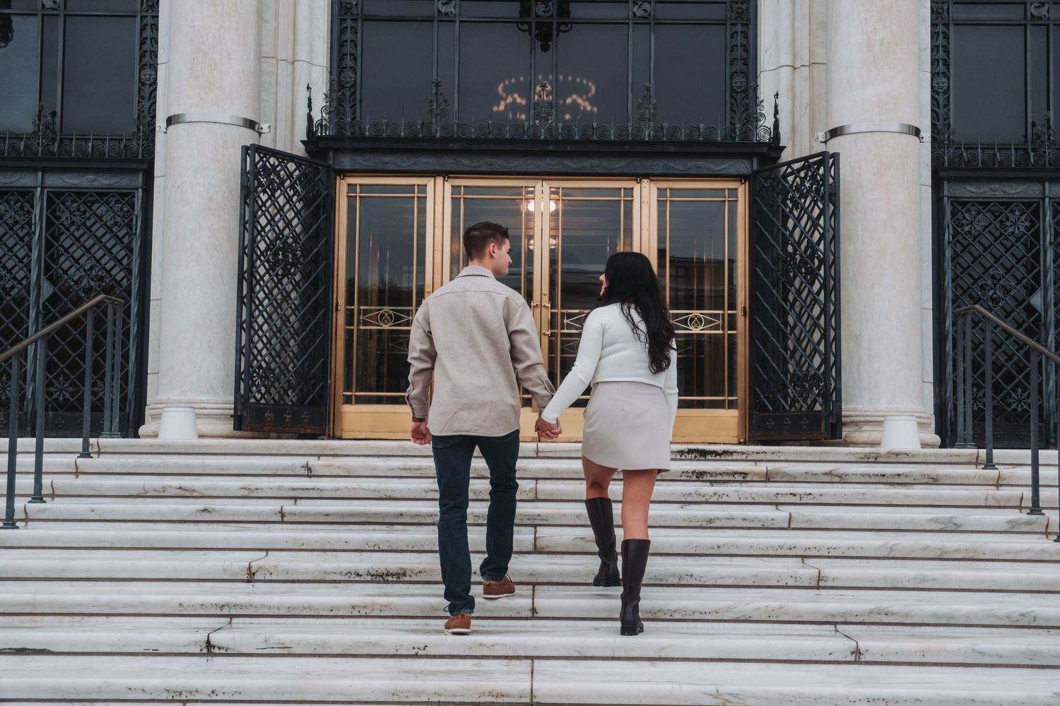 Couple holding hands walking up marble steps toward elegant building entrance with ornate black and gold doors.