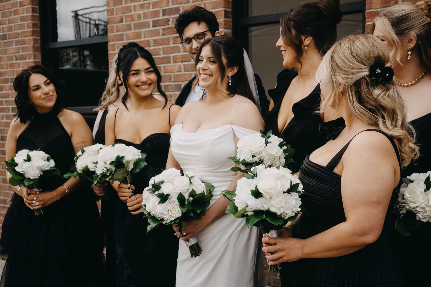 Bride in white gown with bridesmaids in black dresses holding white floral bouquets at an outdoor wedding.