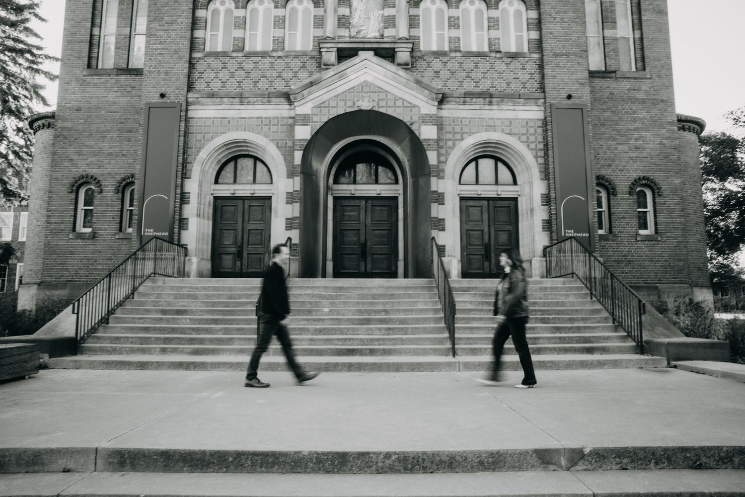 Two people walk in opposite directions on a plaza in front of a historic brick church with arched doorways.
