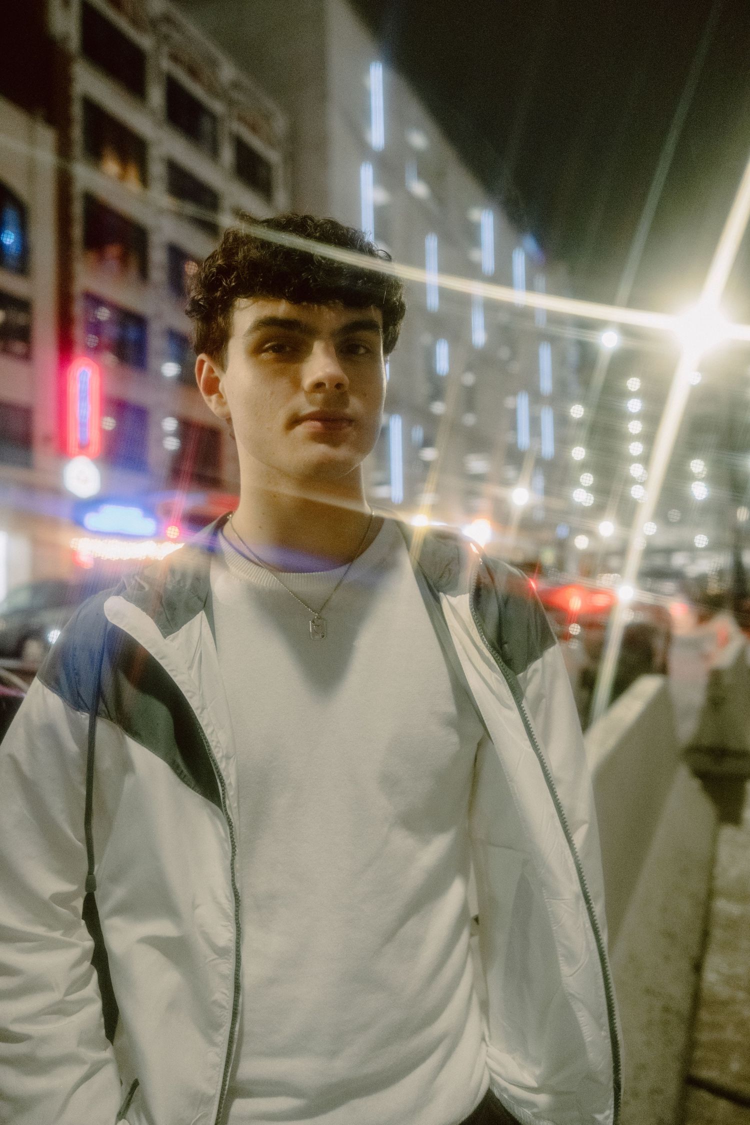 Young man in white jacket standing on a city street at night with neon lights glowing in the background.