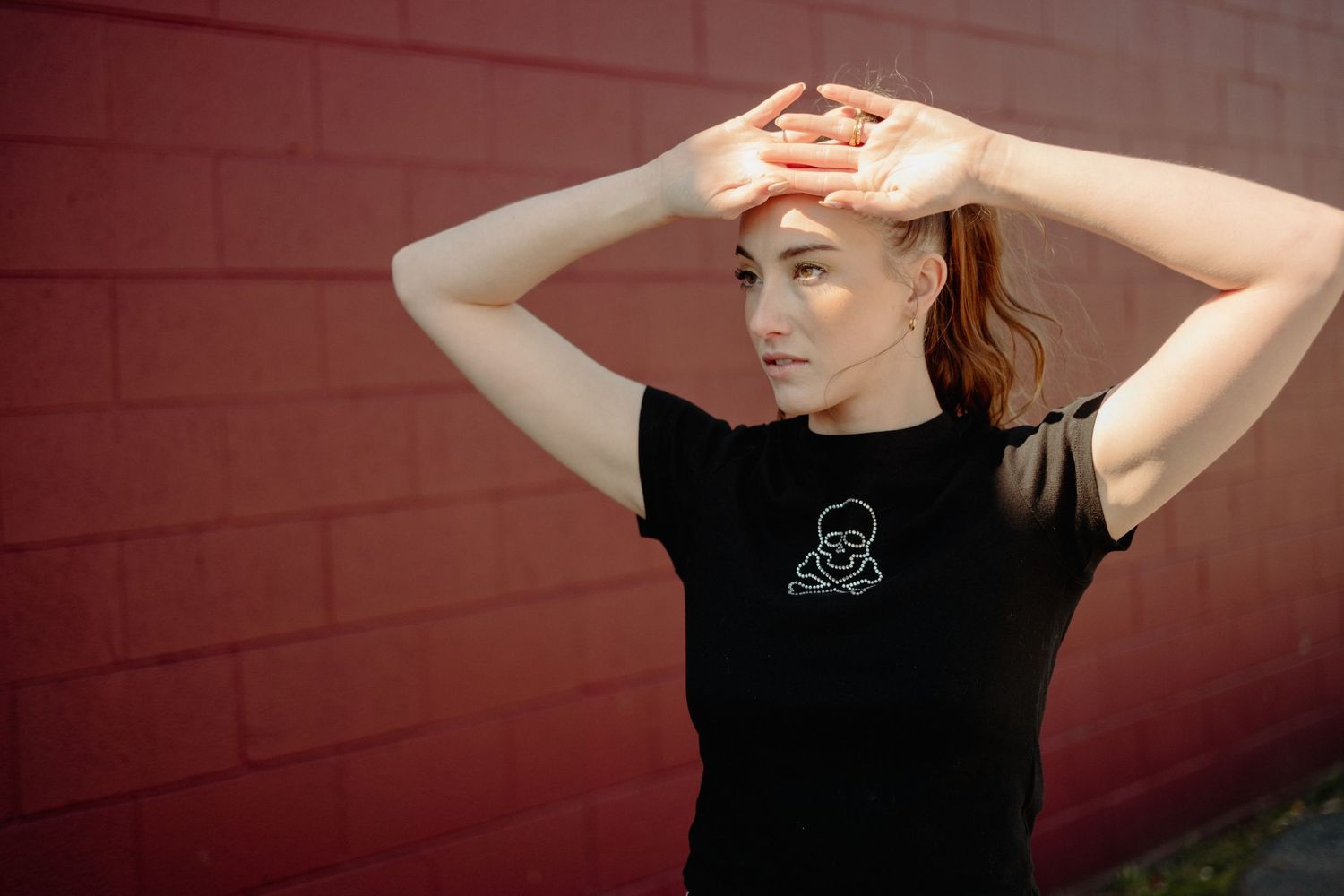 Woman in black graphic t-shirt shielding eyes from sun against red brick wall.