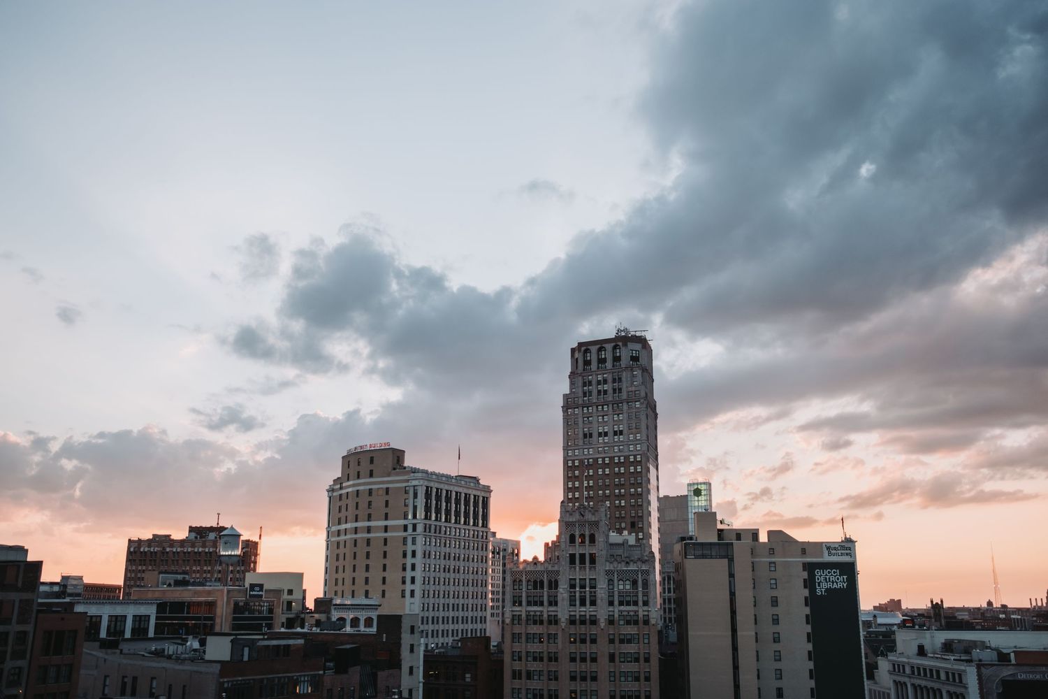 City skyline at dusk with dramatic clouds, featuring a tall historic tower and various buildings bathed in warm sunset light.