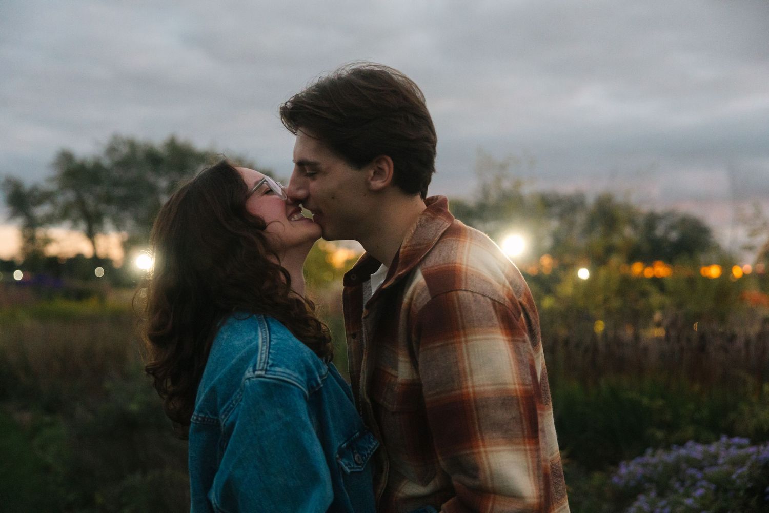A couple in autumn attire stands together against a scenic outdoor backdrop at dusk.