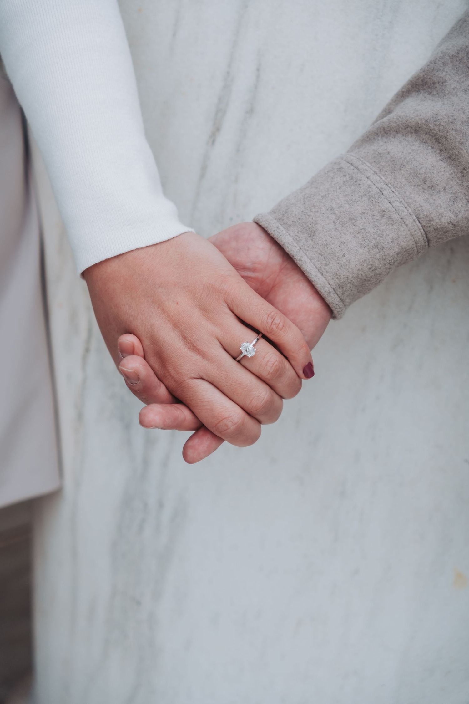 Close-up of two people holding hands, one wearing a diamond engagement ring, dressed in white and gray outfits.