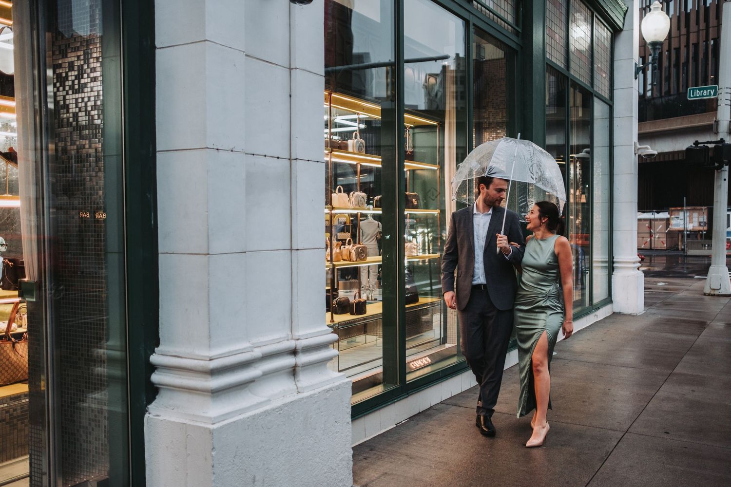 Couple walking under a clear umbrella past a storefront on a rainy city sidewalk, dressed in elegant attire.