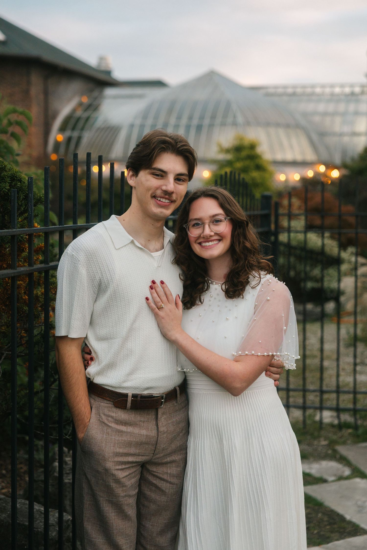 A smiling couple poses together outdoors near a greenhouse, woman in white dress and man in cream shirt and tan trousers.