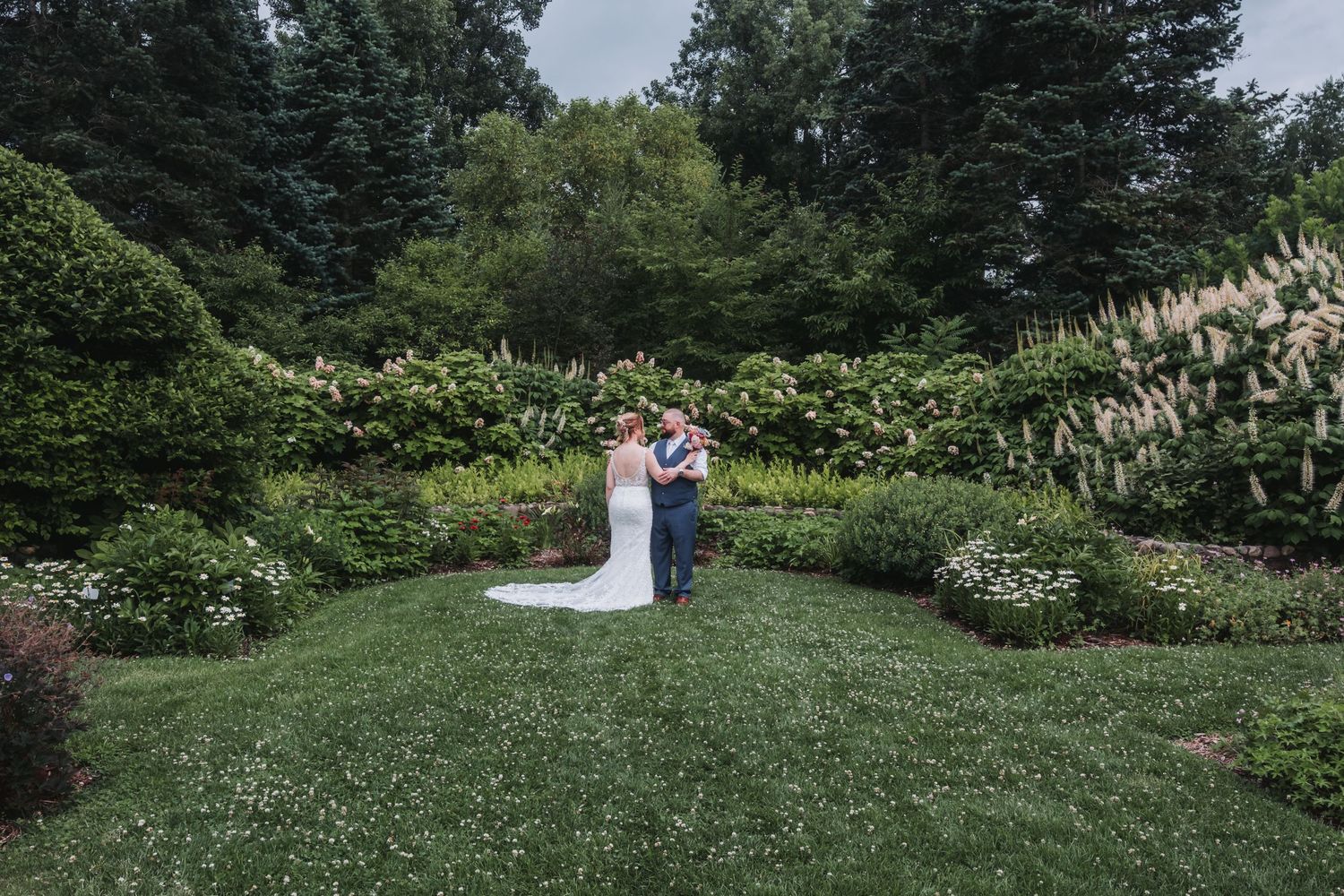 Panoramic view of a wedding couple on a grassy hillside surrounded by trees and flowers.