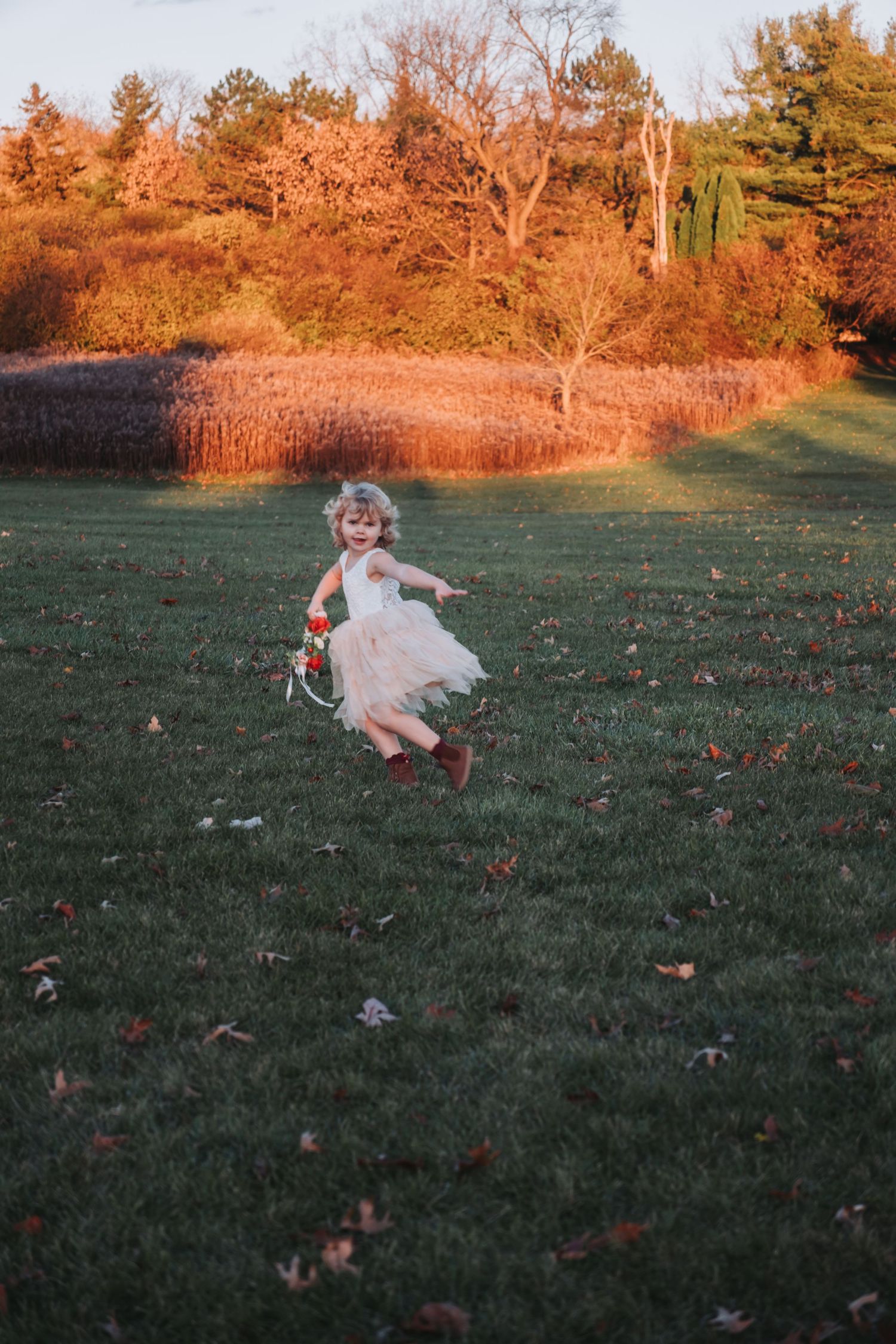 A joyful little girl in a white tutu dress and red boots runs across a green lawn with autumn foliage in the background.