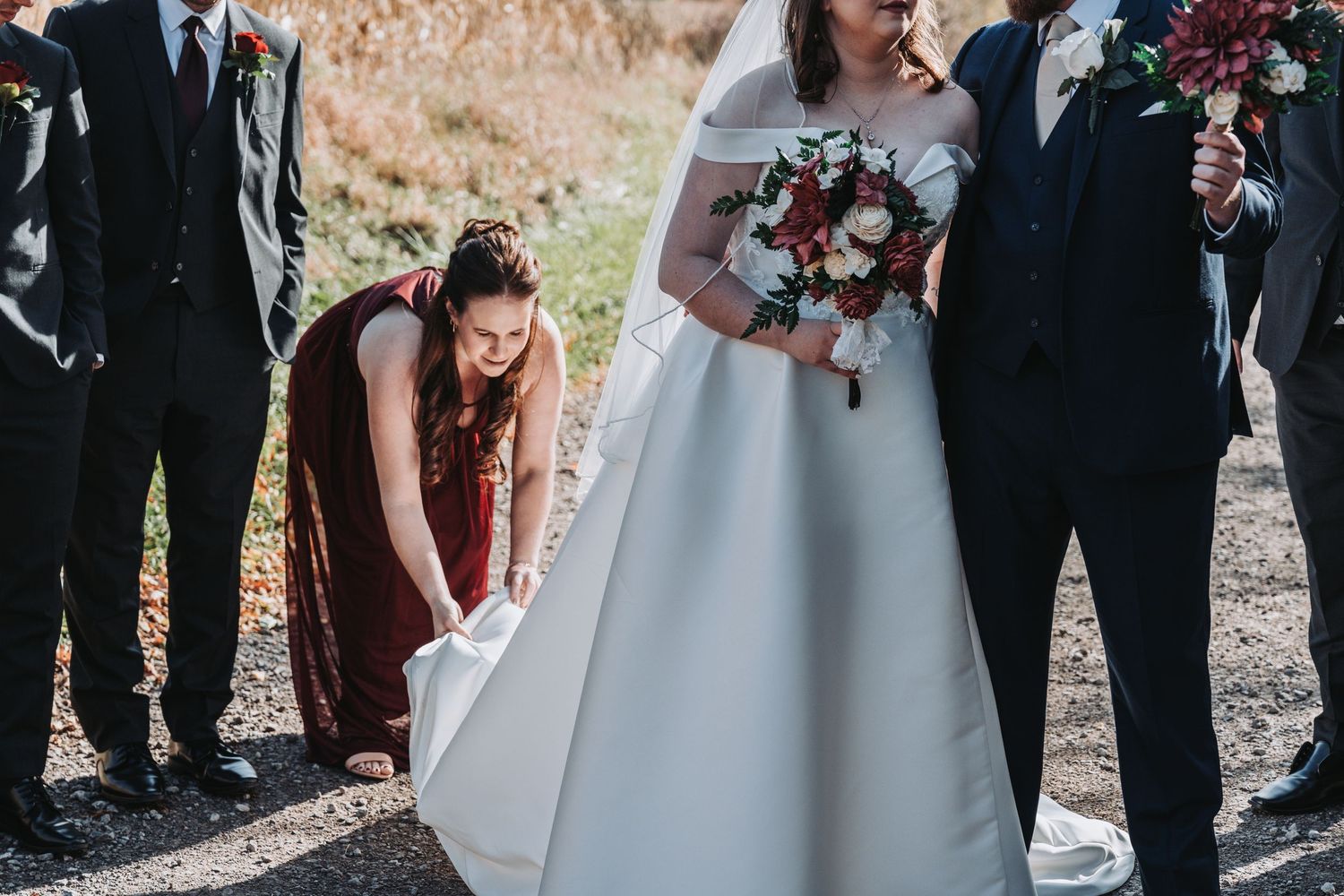 Bridesmaid in burgundy dress adjusting bride's white wedding gown while groomsmen stand nearby holding bouquet.