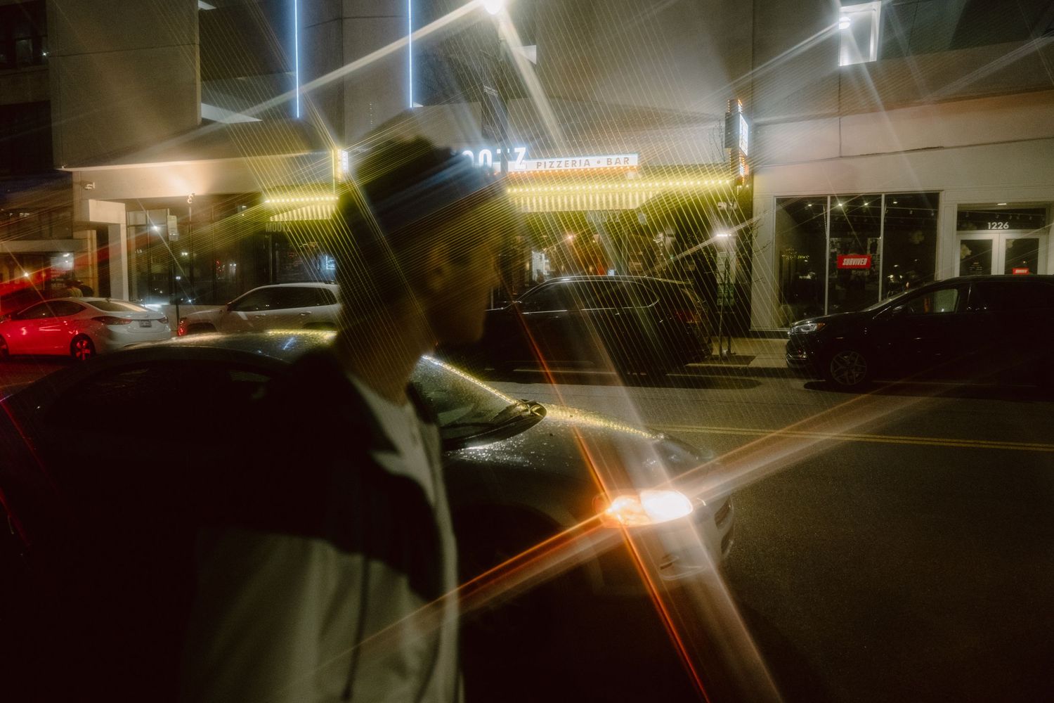 Man in beanie hat standing on urban street at night with colorful light streaks and illuminated storefronts.