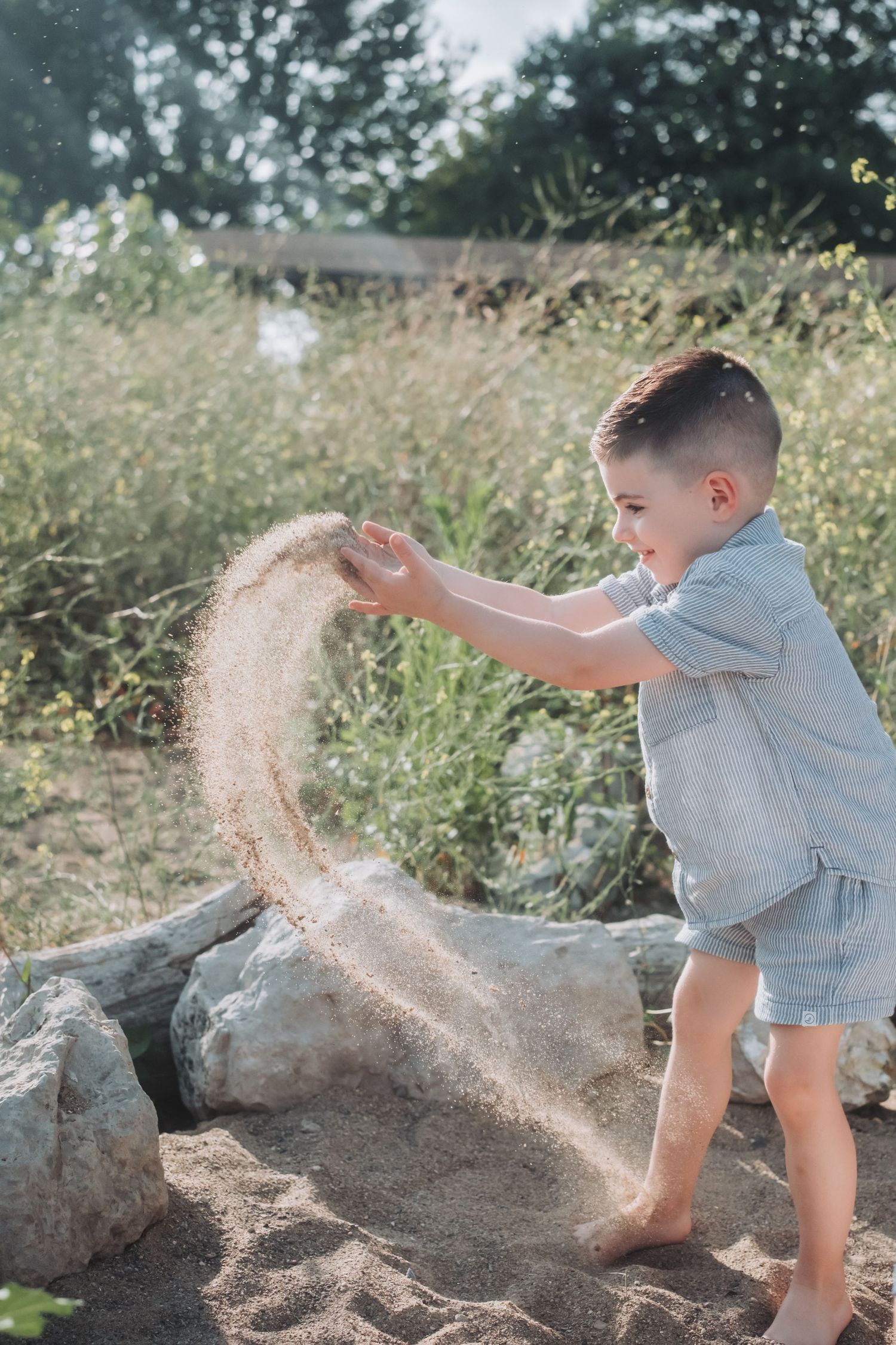 Young boy playing with sand outdoors, letting it fall through his hands near rocks and green vegetation.