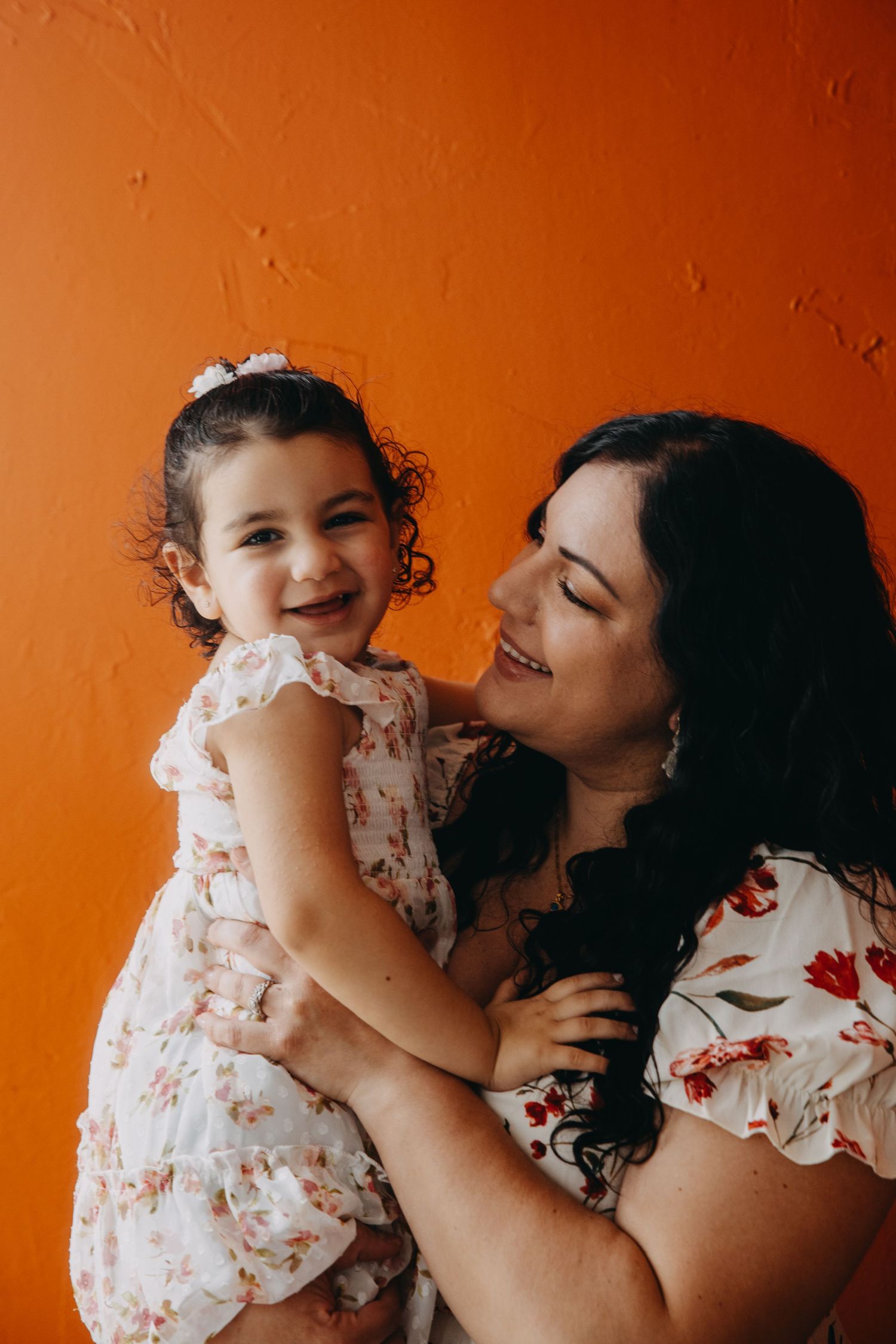 A happy mother holds her laughing toddler daughter, both wearing floral dresses against a vibrant orange wall.