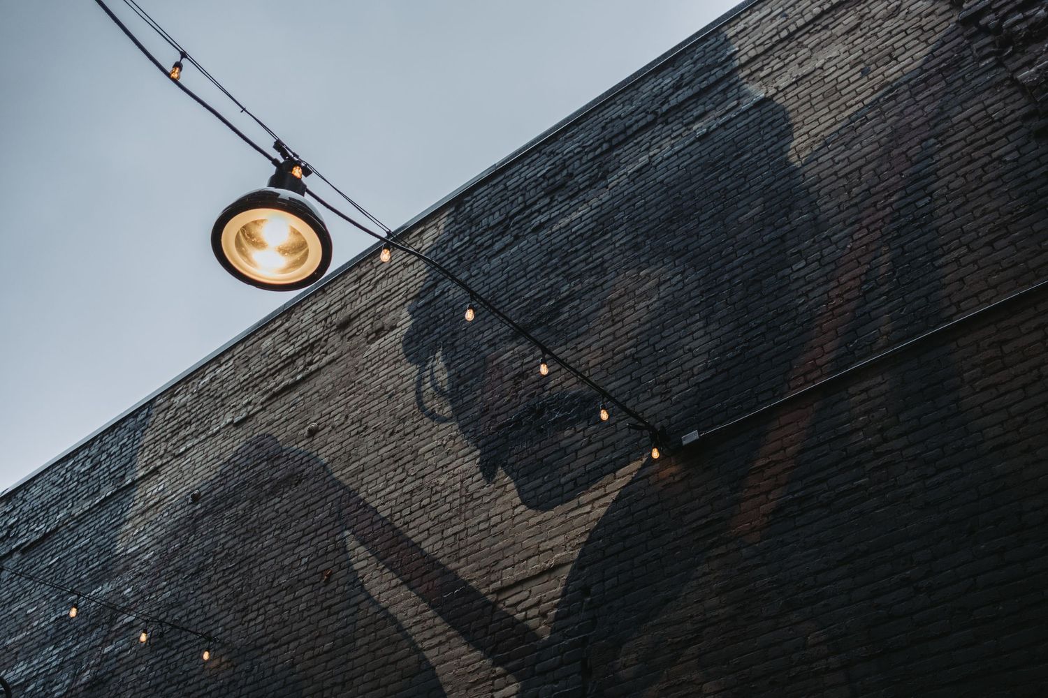 Dark brick building exterior with string lights and a glowing lamp against a cloudy sky, featuring a large mural.