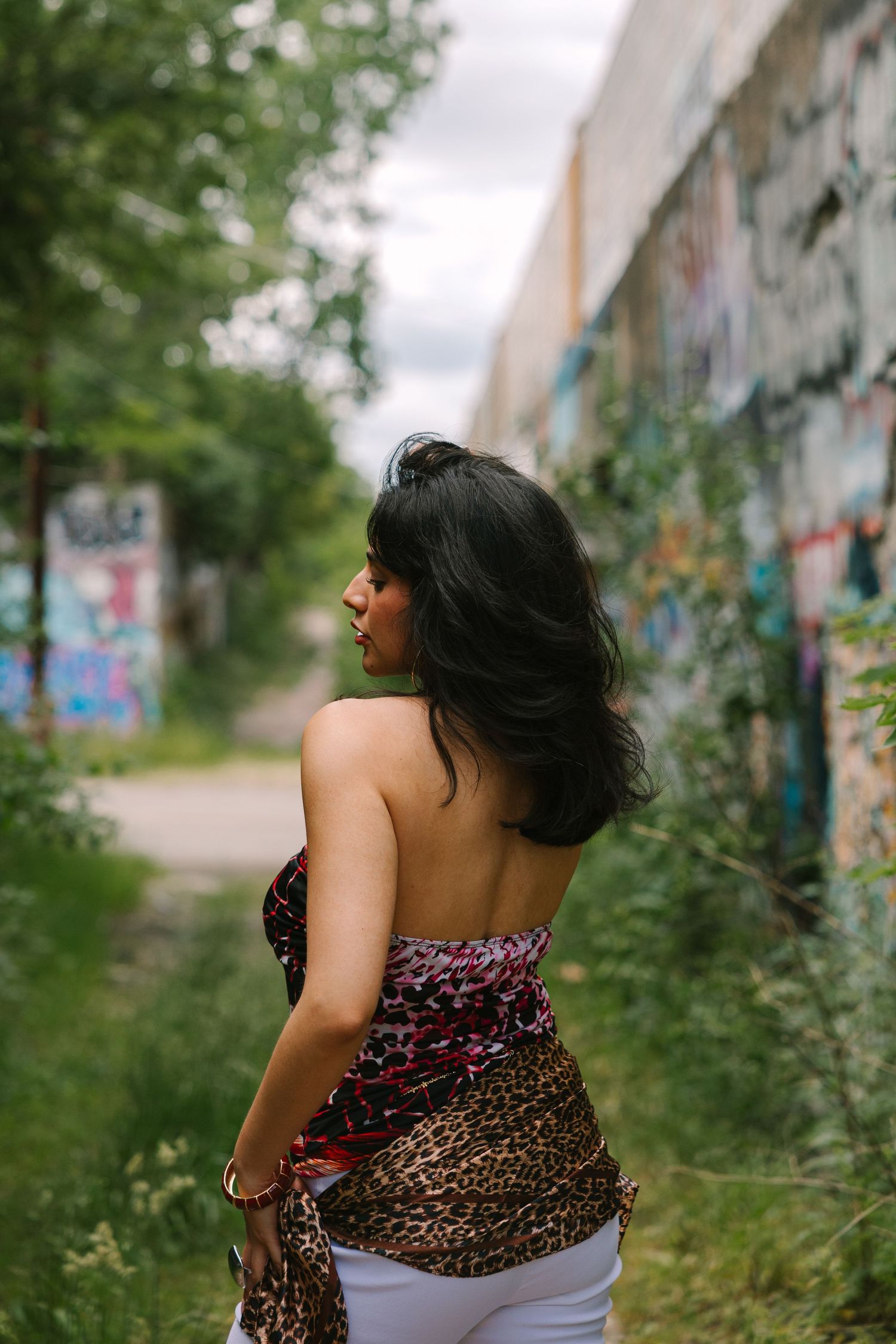 Woman in leopard print halter top with flowing dark hair poses near graffiti wall surrounded by lush greenery.