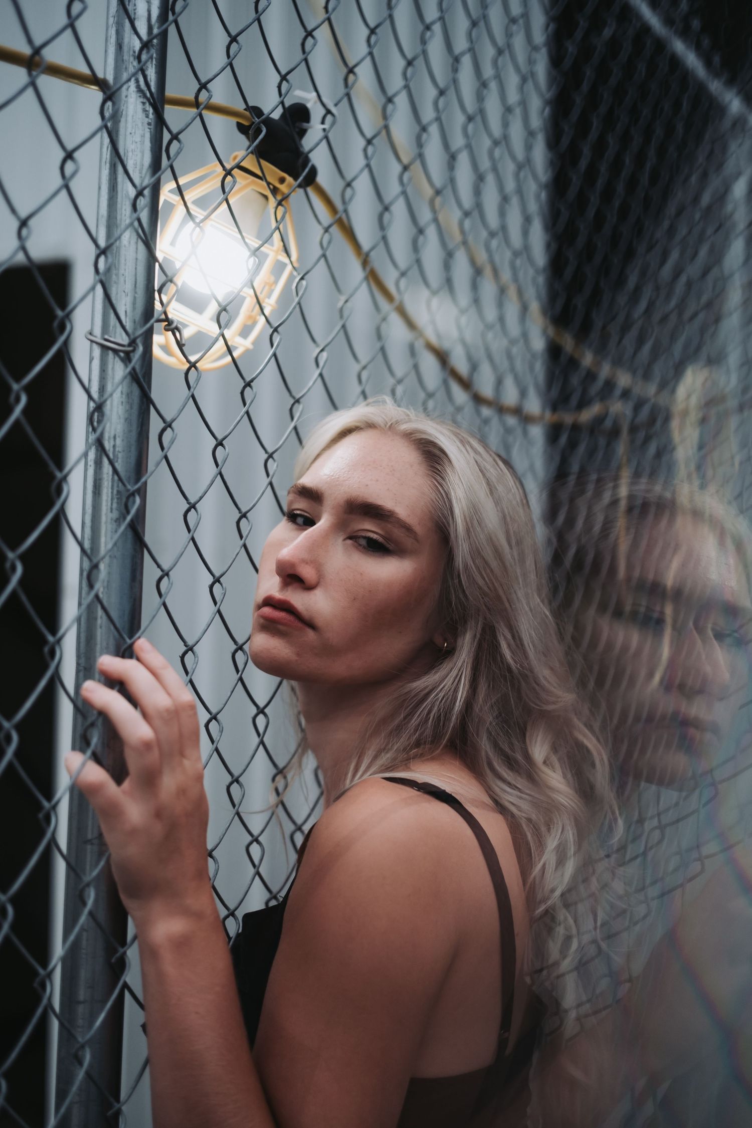 Blonde woman in black top leaning against chain-link fence with glowing bulb light, moody urban portrait.