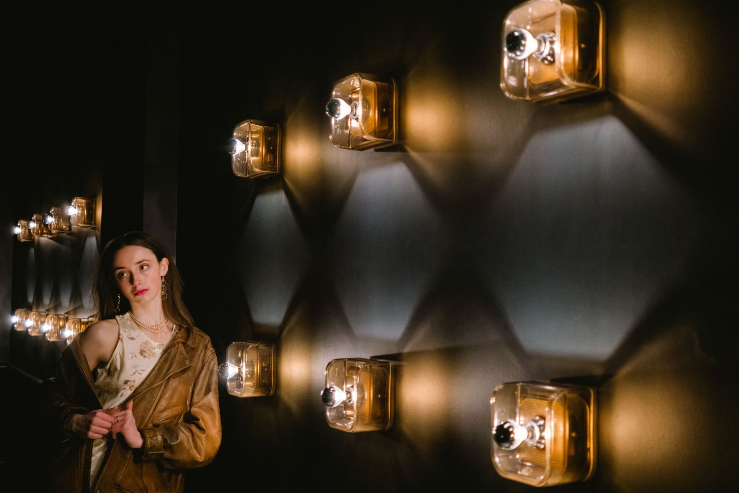 Woman in golden jacket posing beside a dark wall with glowing amber hexagonal light fixtures.