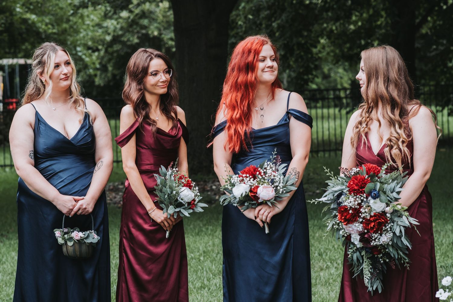Bridesmaids in navy and burgundy dresses holding floral bouquets outdoors.