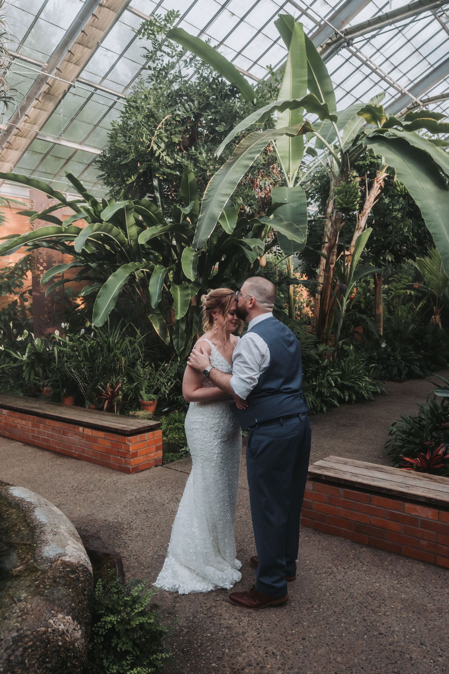A romantic wedding moment captured in a greenhouse surrounded by tropical plants and natural light.