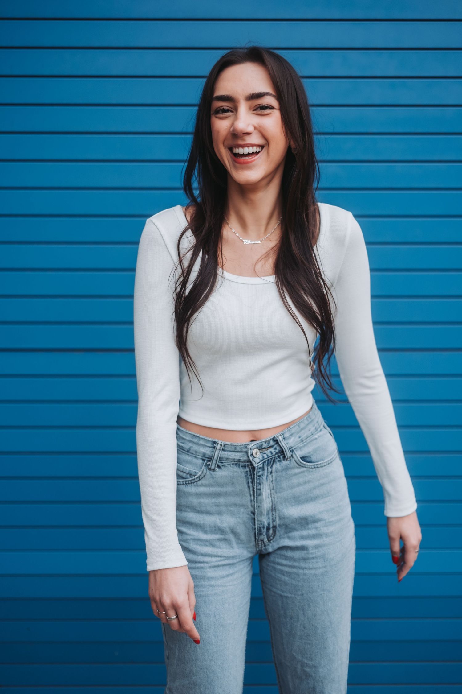 Person in white crop top and light jeans poses playfully against bright blue wall.