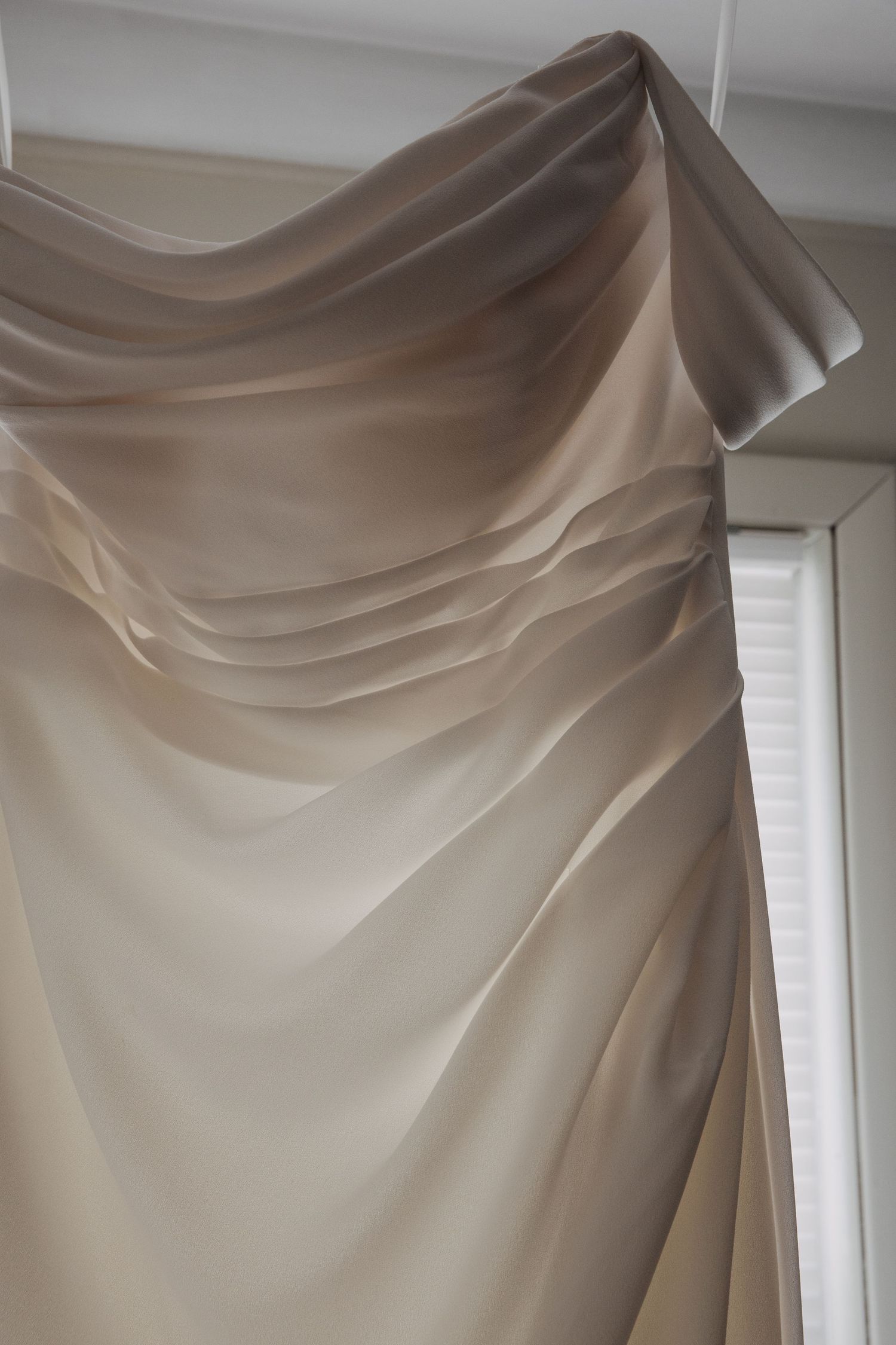 Elegant draped taupe satin fabric cascading in soft folds near a bright window, creating luxurious flowing curtain effect.