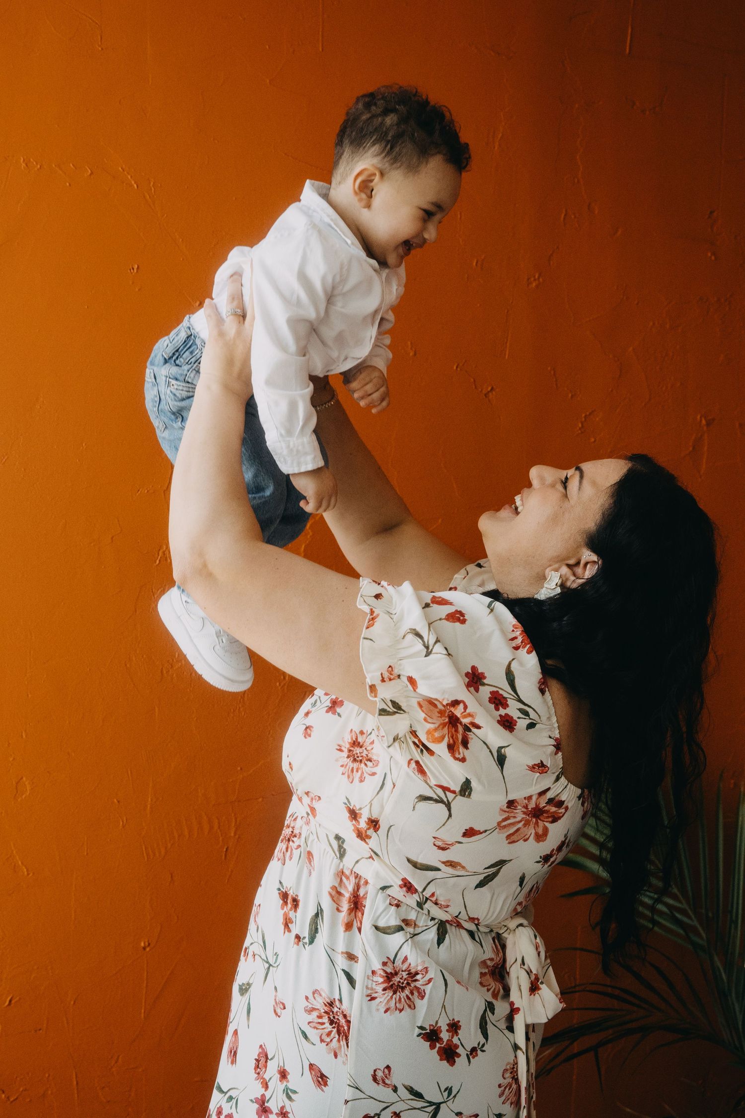 A mother in a floral dress joyfully lifts her smiling baby boy against a vibrant orange wall.