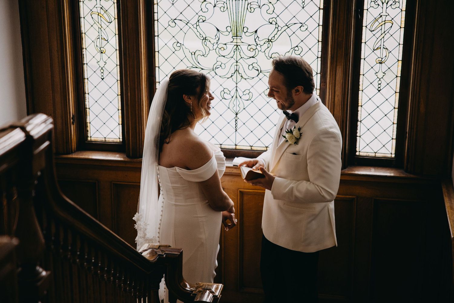 Bride and groom exchange vows by stained glass windows in elegant wood-paneled venue during intimate wedding ceremony.