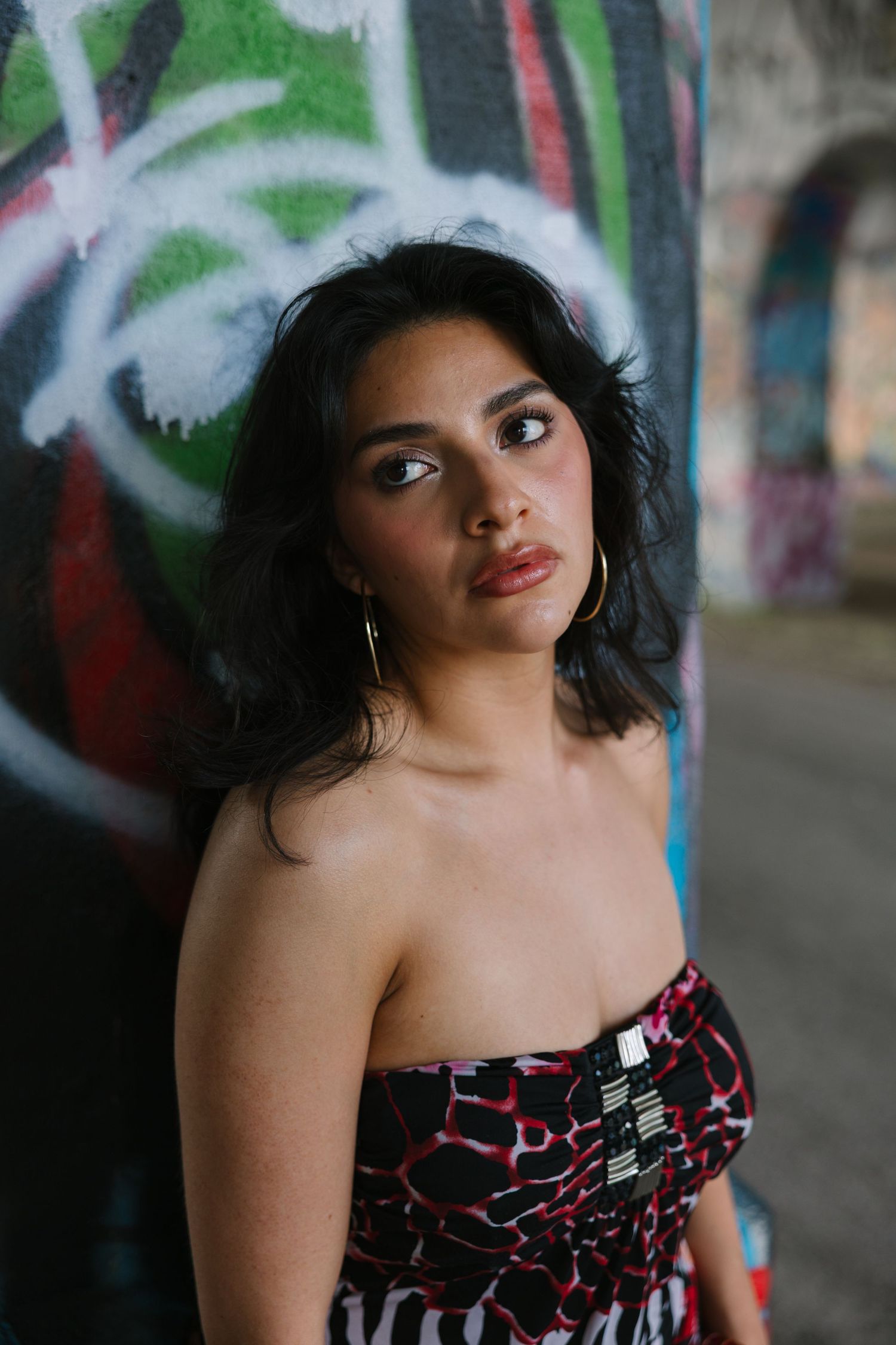 Young woman in strapless patterned dress poses against colorful graffiti wall, wearing hoop earrings.