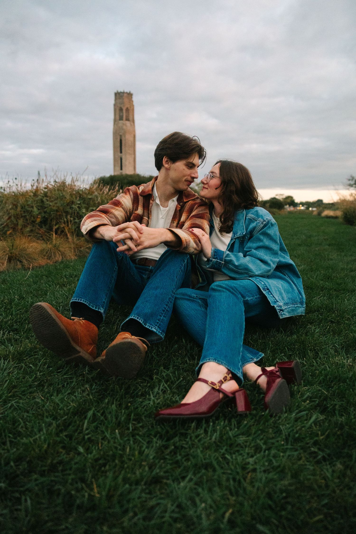 Romantic couple leaning in for a kiss while sitting on grass with a stone tower backdrop.