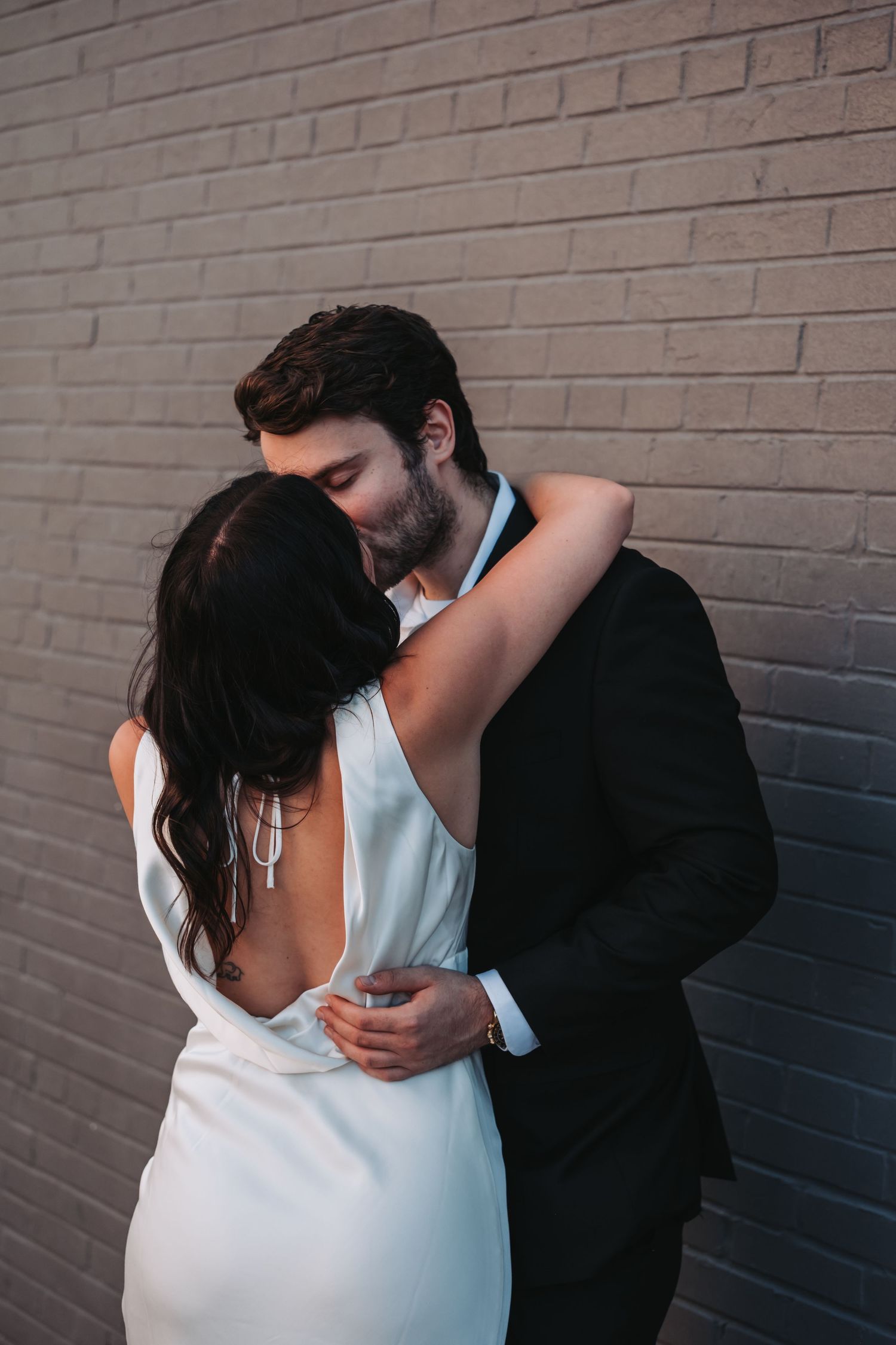 Couple sharing a romantic kiss against a brick wall, bride in backless white gown, groom in black suit.