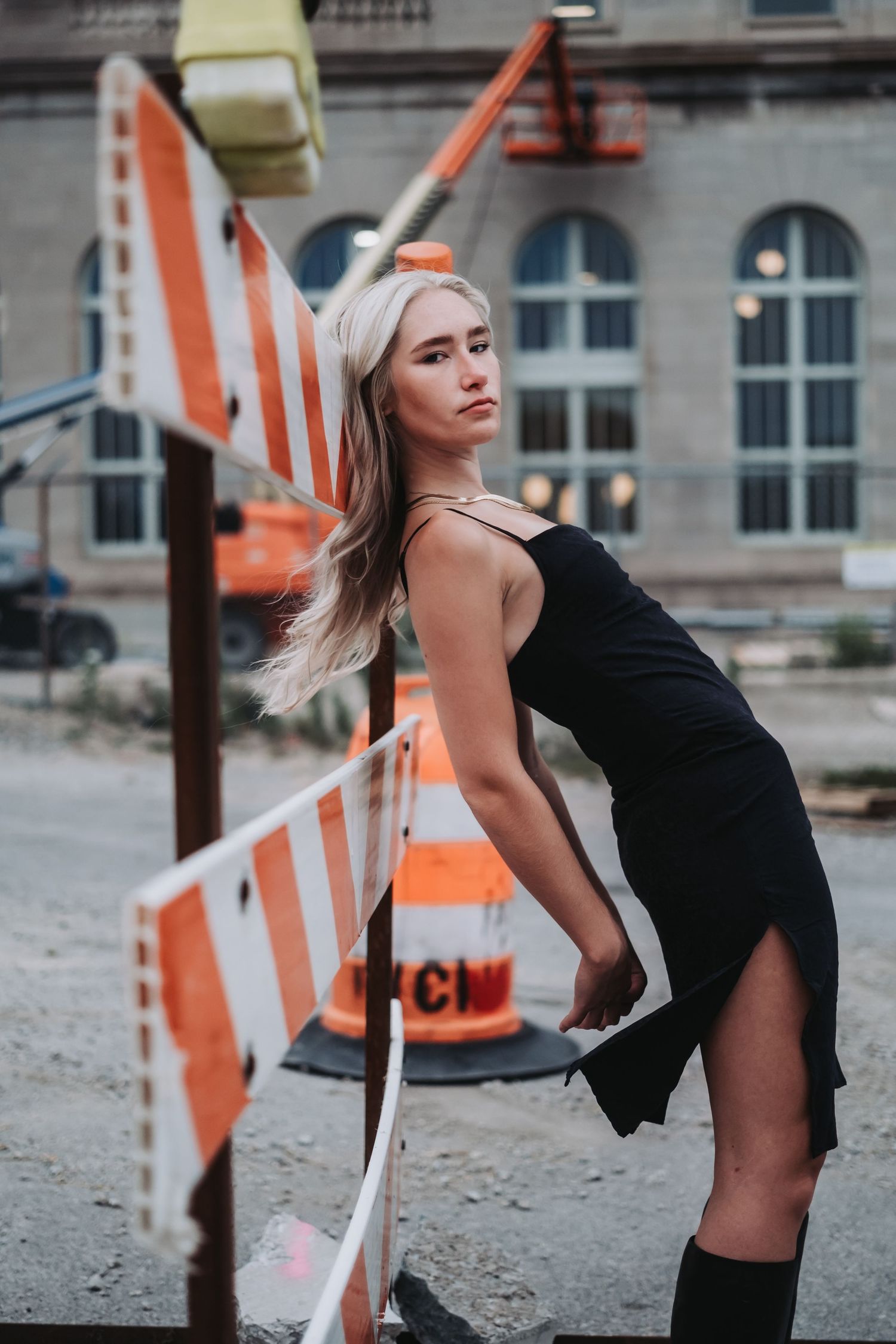A woman in black poses dramatically near orange construction barriers in an urban setting.