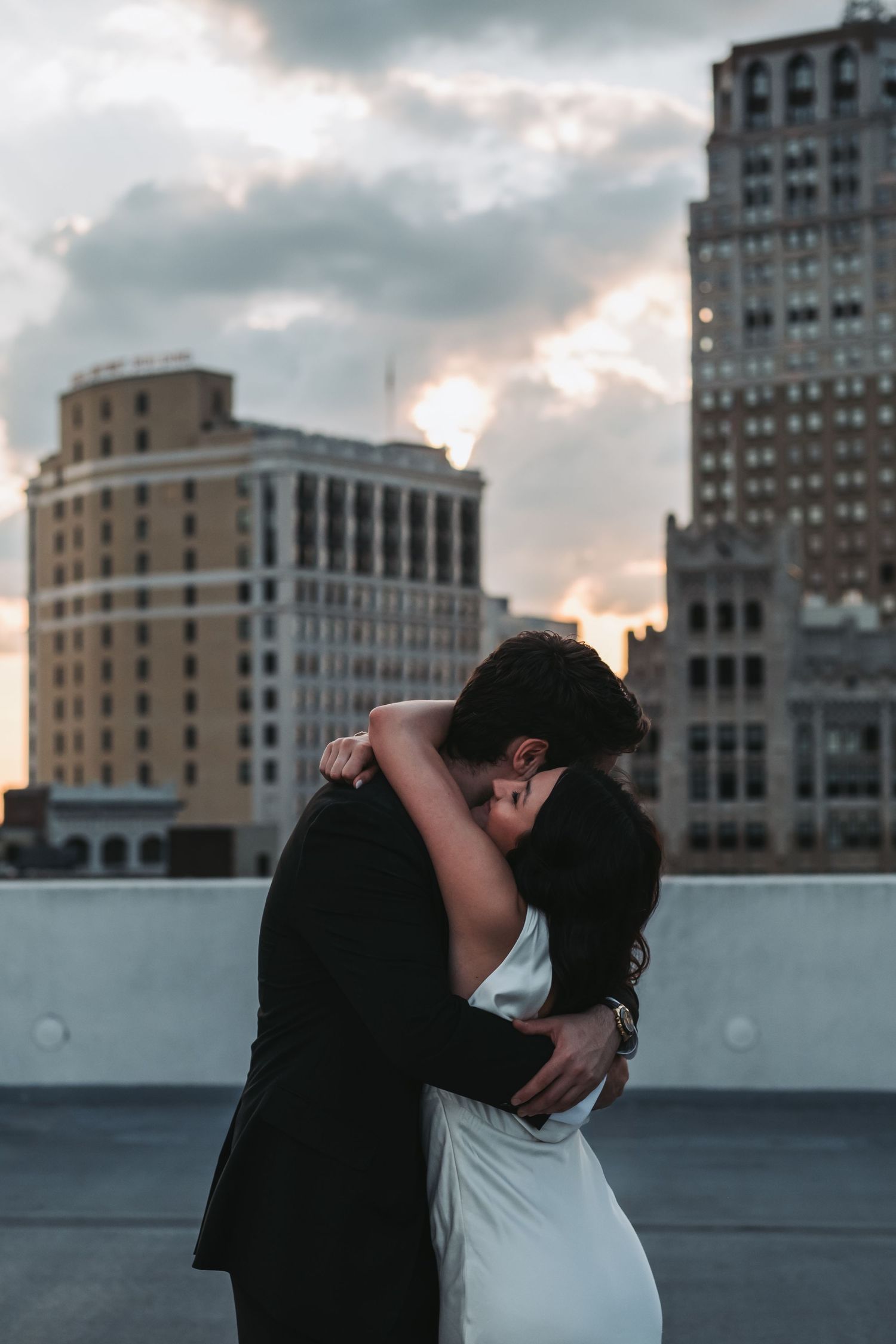 A couple in wedding attire share a passionate kiss on a rooftop at sunset with a stunning city skyline backdrop.
