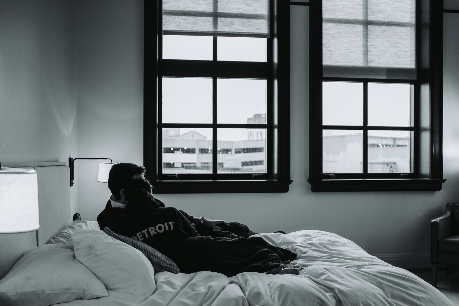 Person in Detroit hoodie sitting alone on bed in monochrome bedroom with large windows and city view.