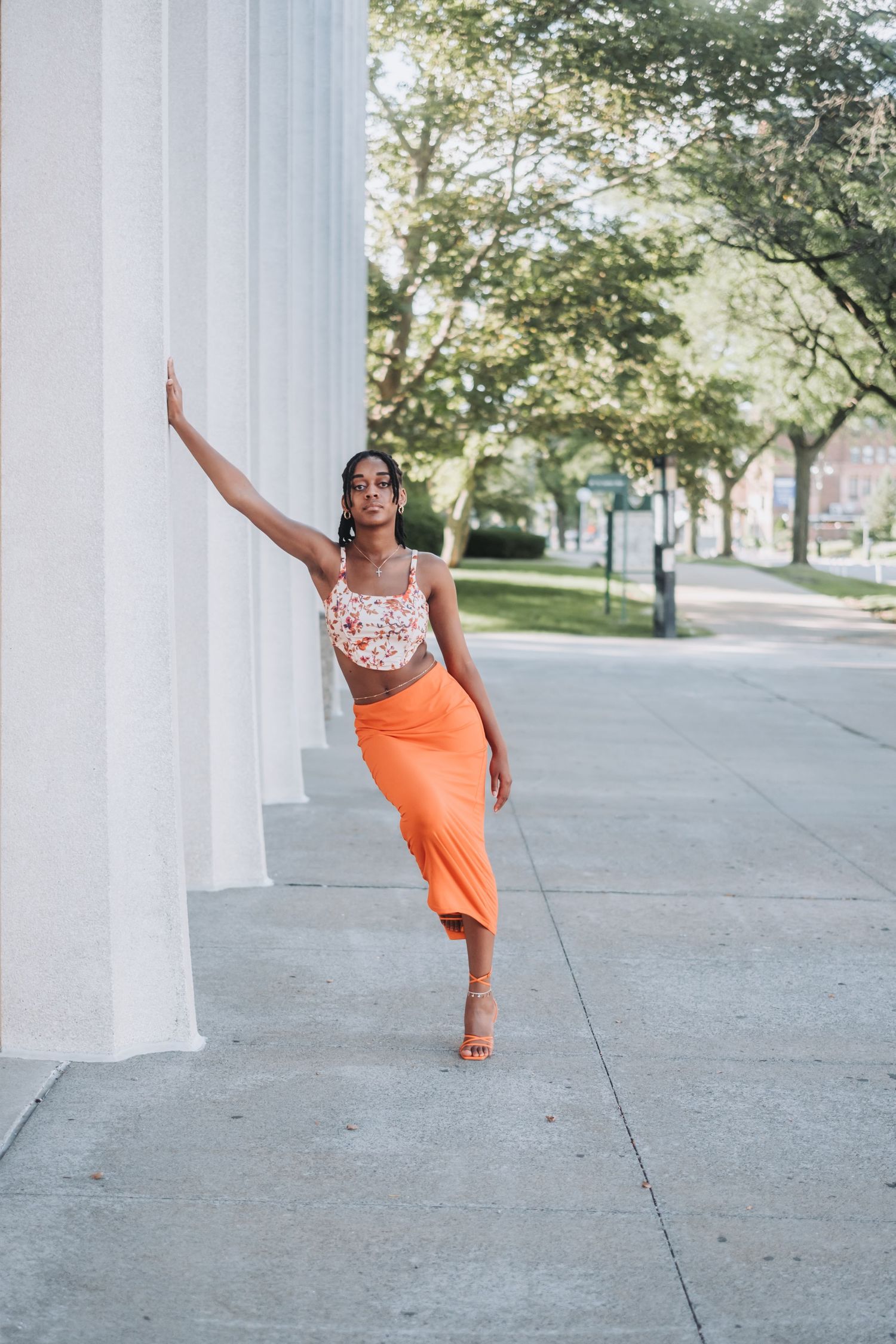 A woman in an orange midi skirt and floral crop top poses gracefully against a white column on a sunny sidewalk.