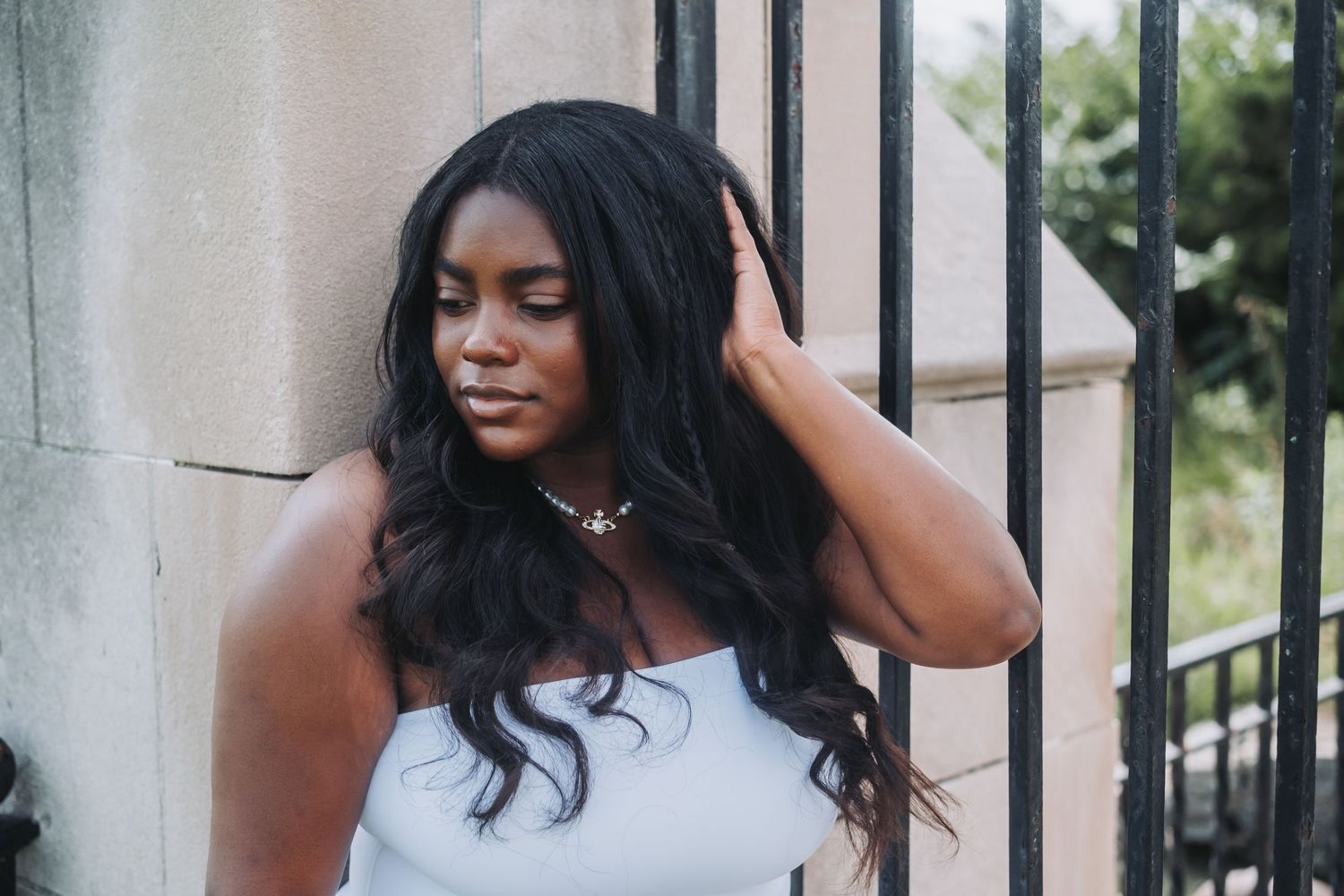 Young Black woman with long wavy hair poses gracefully near iron gate, wearing white top and gold necklace outdoors.