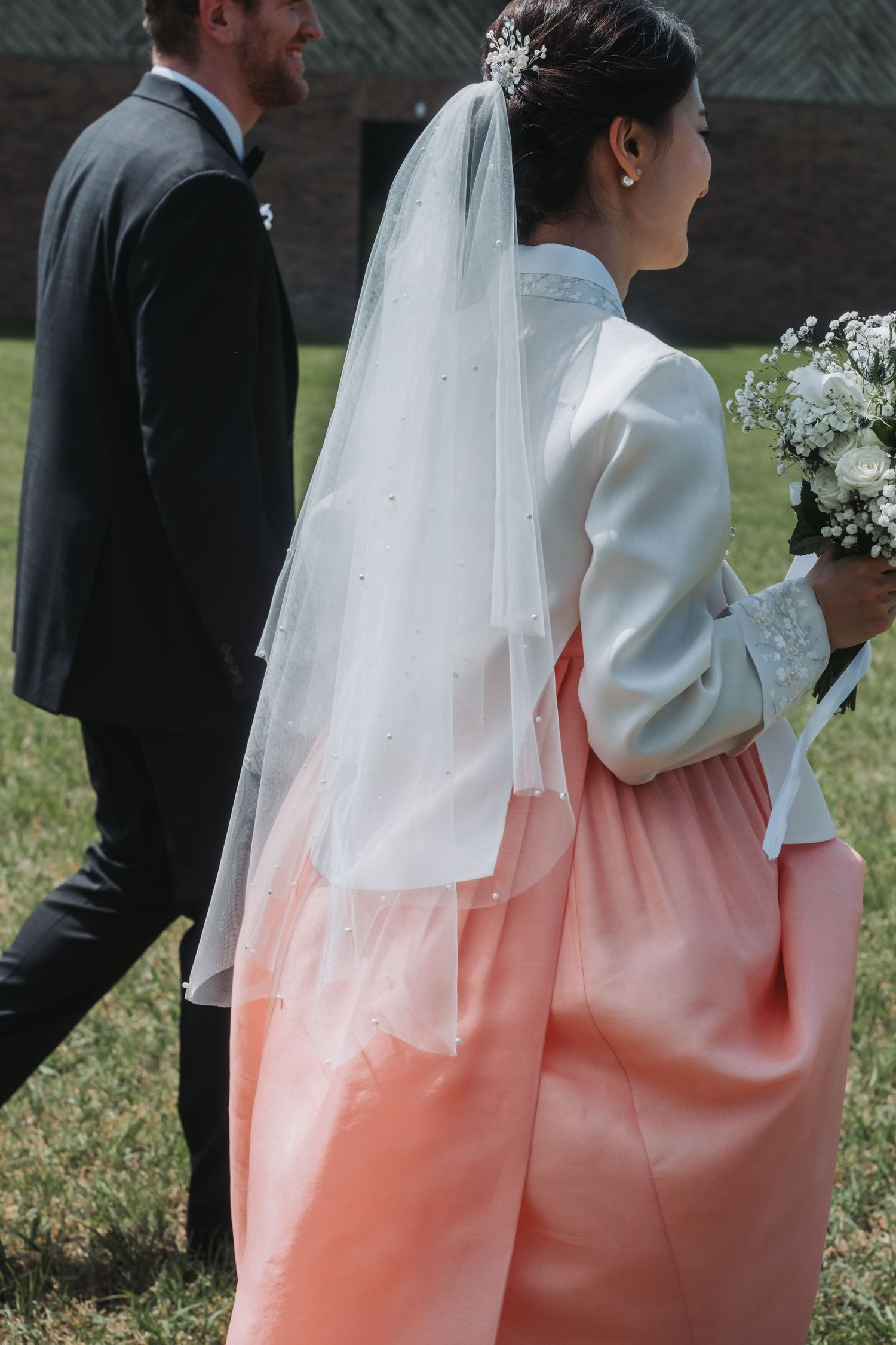 Bride in traditional white wedding dress with flowing veil walks outdoors holding a bouquet.
