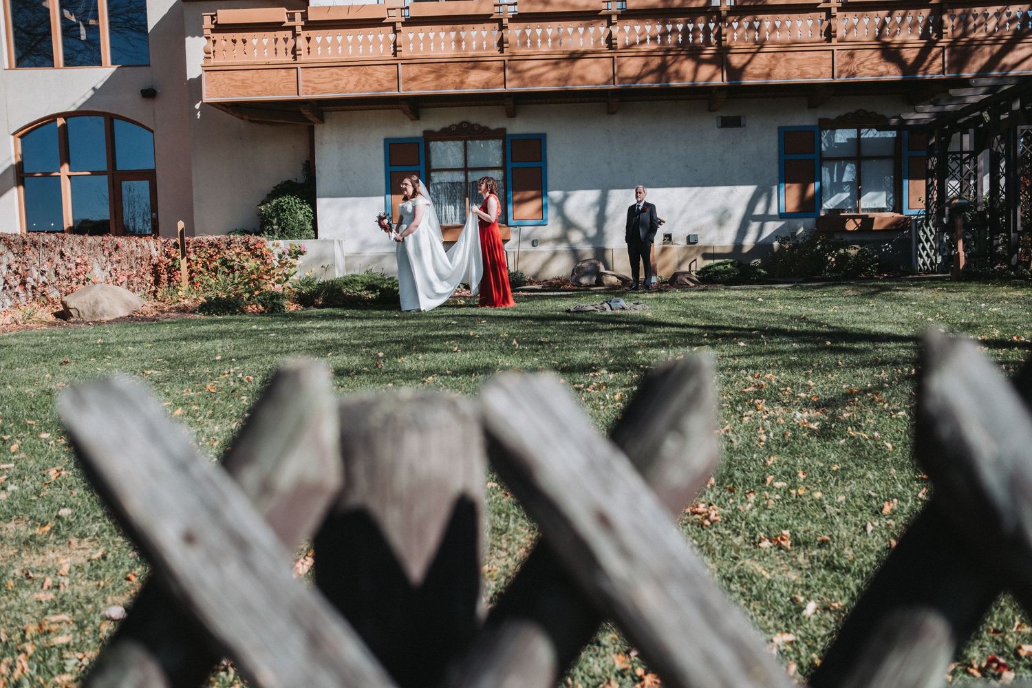 A wedding party gathers on a sunny lawn viewed through a wooden fence.